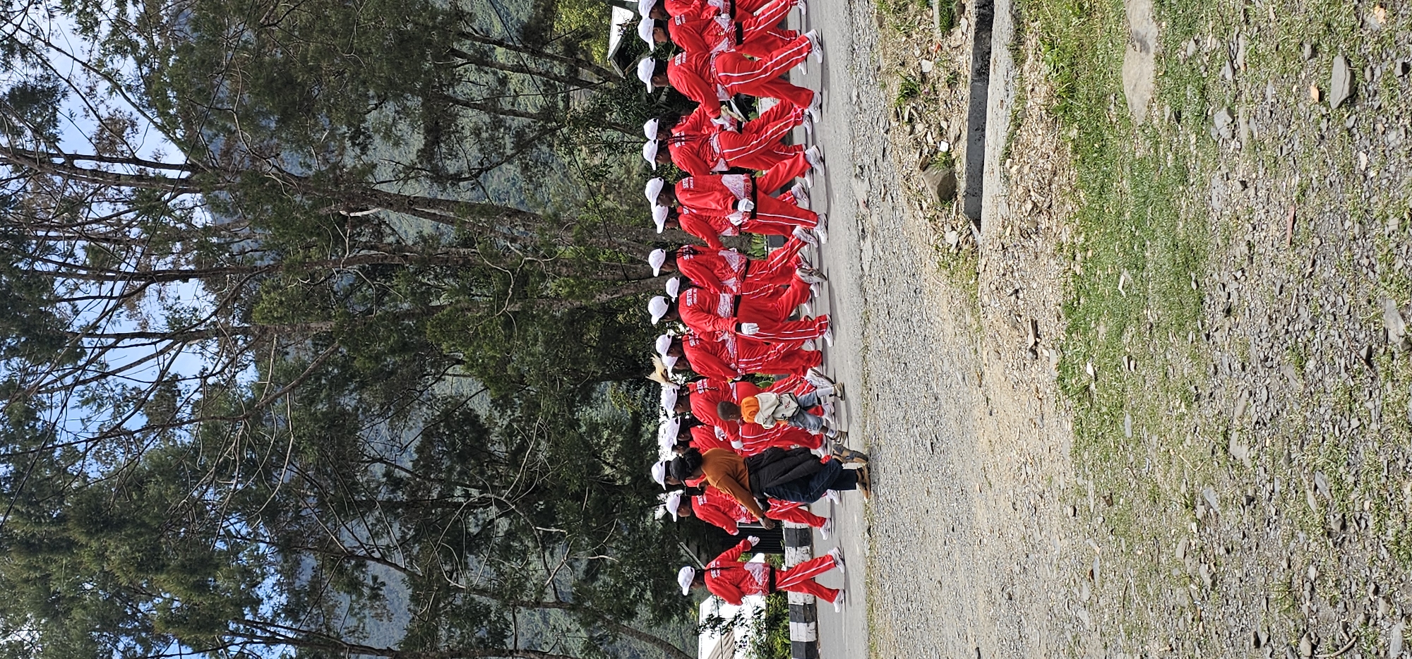 In Puncak Jaya, Indonesia, a group of young people in matching bright red tracksuits and white caps are marching in formation on a paved road. The group appears to be a marching band or a similar uniformed organization, with many members visible in the frame. A woman in an orange shirt and dark pants is walking alongside the formation, and a small child in an orange hooded jacket is also present. The environment consists of a tree-lined road with lush green hills and vegetation visible in the background. The sky is overcast, and the overall atmosphere suggests an outdoor event or parade.