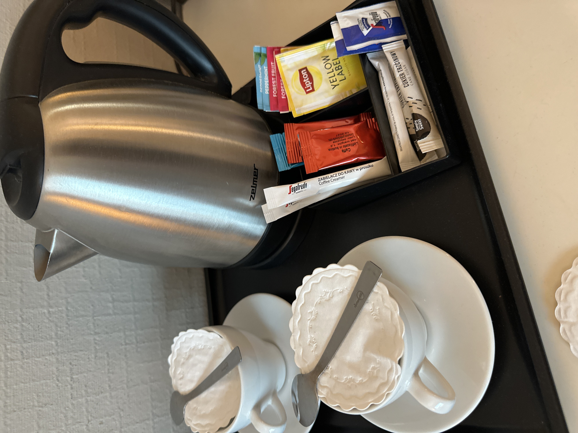 A close-up shot depicts a refreshment station on a black tray, containing a Zelmer electric kettle, two cups with spoons and decorative paper coasters, and a selection of coffee and tea sachets.
The kettle, silver and black, is positioned on the left side of the frame. Two white cups with saucers and silver spoons lie on the tray, covered with decorative paper coasters.
A small black container holds an assortment of tea and coffee sachets, including brands like Lipton Yellow Label, Forest Fruit tea, Segafredo coffee creamer, and Simply Sweet cane sugar.
While the image indicates a hotel room refreshment station, there is no information within the image itself that specifies the location of this room as Wrocław, Poland.
