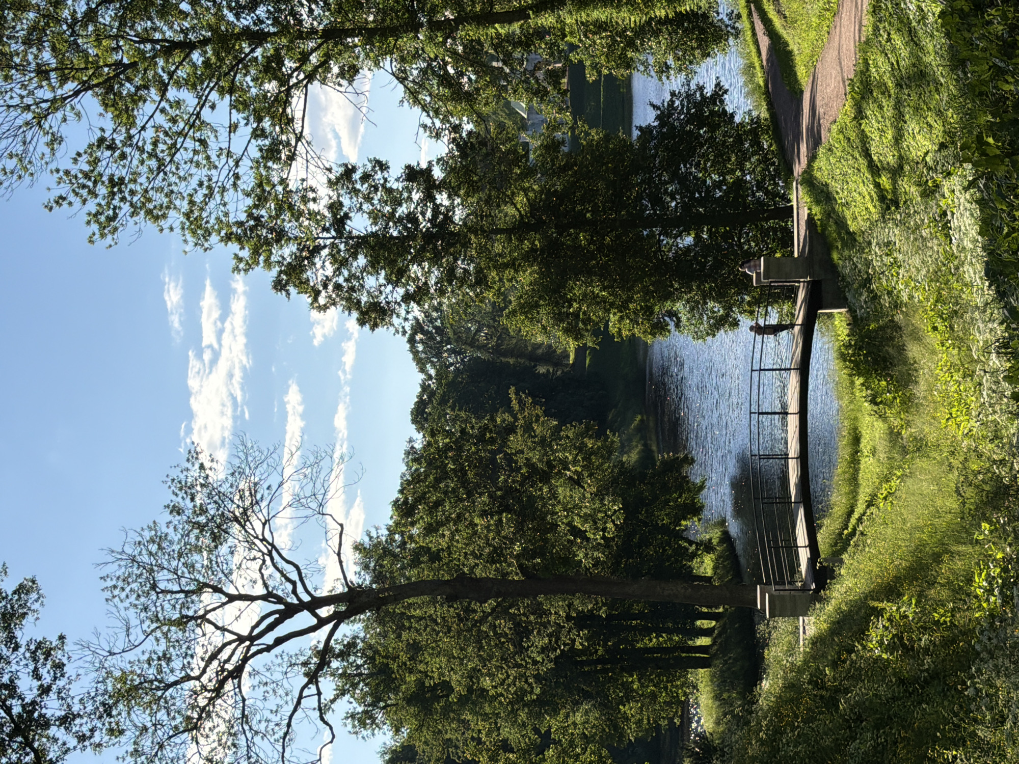 In an outdoor scene under a partly cloudy blue sky, a small footbridge spans a body of water. The bridge has a dark railing supported by two stone pillars, one of which has a light-colored cap. A person stands on the bridge, facing away from the camera and towards the water. The surrounding environment is lush with green trees and vegetation. To the right of the bridge, a dirt path winds uphill, also bordered by green grass. In the background, across the water, distant trees and a building with a light-colored facade and green roof are visible. The sun casts shadows from the trees onto the grass and water, indicating it is likely late afternoon.