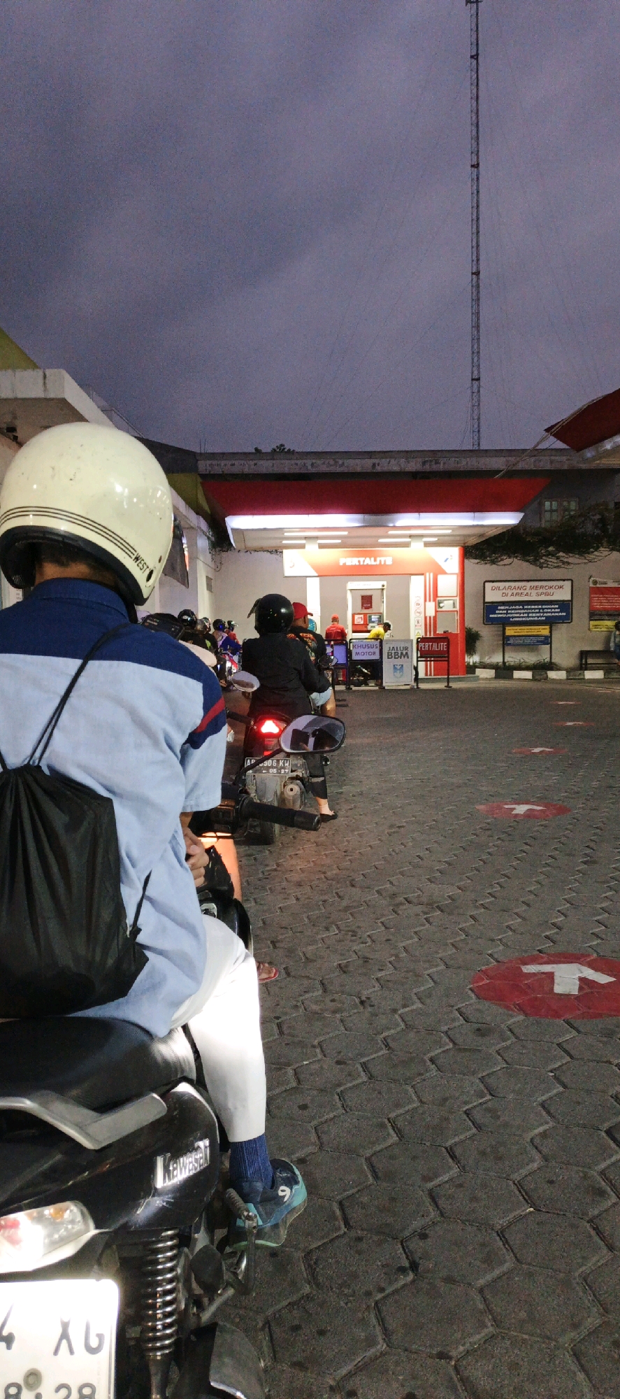 In Kasihan, Indonesia, at dusk, a long queue of motorcyclists waits at a Pertamina gas station. The sky is a muted purple, and a tall communication tower stands in the background. The station features a red and white awning covering the pumps, with signs indicating "Pertalite" fuel and directives for motorcyclists. Red and white arrows on the ground mark the queue. The foreground shows a motorcyclist from behind, wearing a white helmet and a blue and white shirt, with a black bag slung over their shoulder. Another motorcyclist in front wears a black helmet and red shirt. The overall atmosphere is one of routine activity at a public service point.