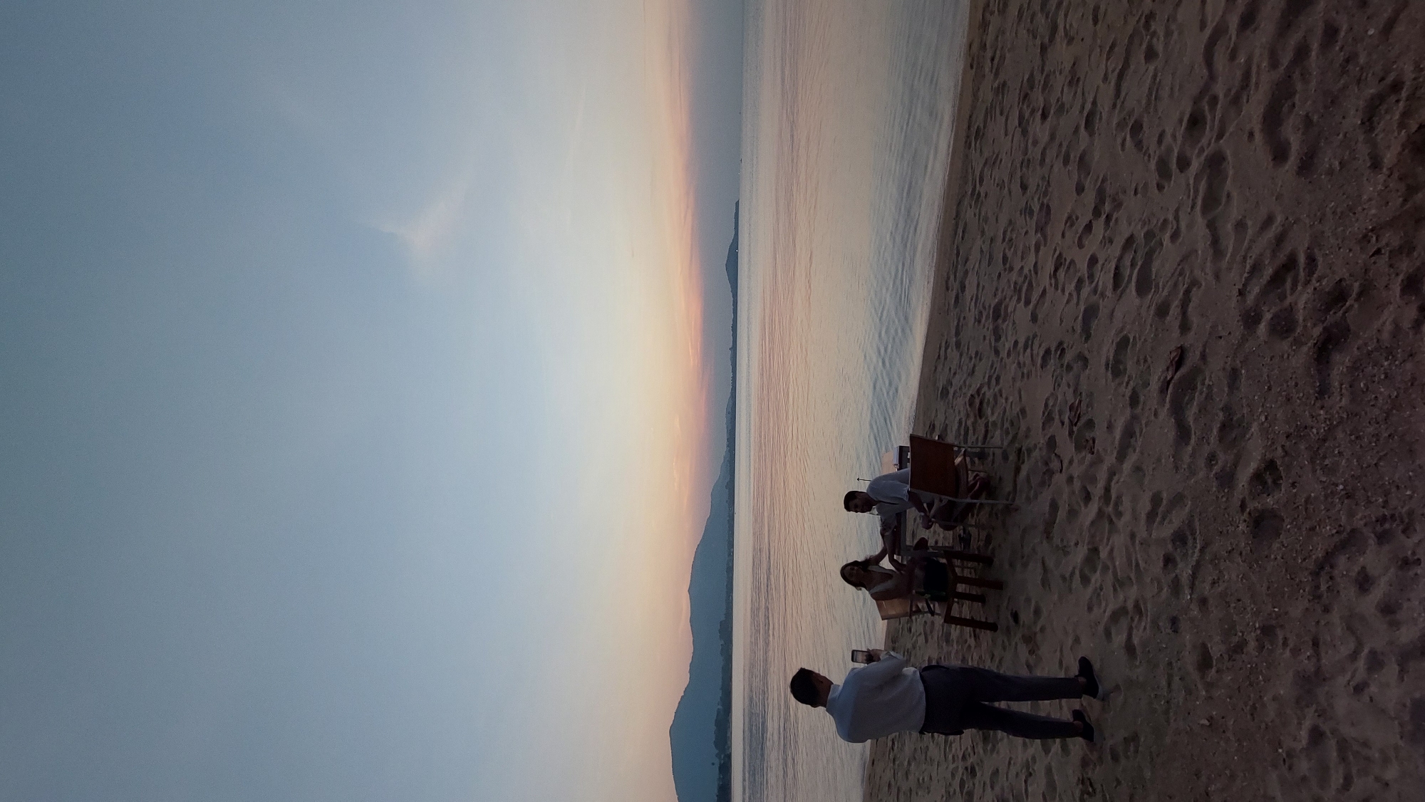 Baan Bo Phut, Thailand. A sunset scene at a beach. Three people sit at a table and chairs near the shoreline. Another person stands taking a photo of the group with their phone. The beach sand shows numerous footprints. The sky is a gradient of blue to orange. Mountains are visible across the water.