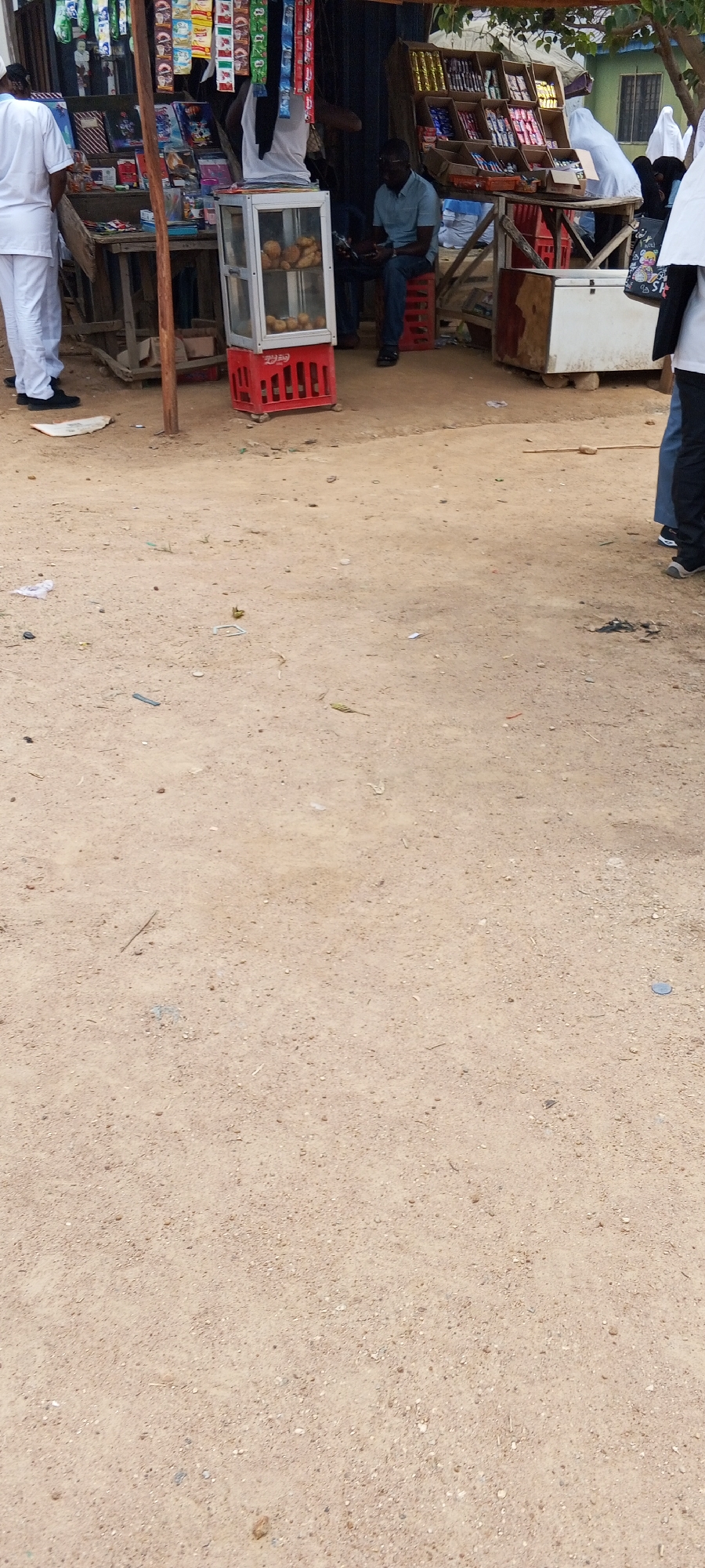 The image shows a street scene in Keffi, Nigeria. A small roadside shop is visible, constructed from wood and metal, with a display of various goods including snacks, books, and baked goods in a glass display case. A man is seated on a red crate near the shop, appearing to be using a mobile device. A few other people are standing near the shop, some wearing white clothing. The ground is dirt and sand.