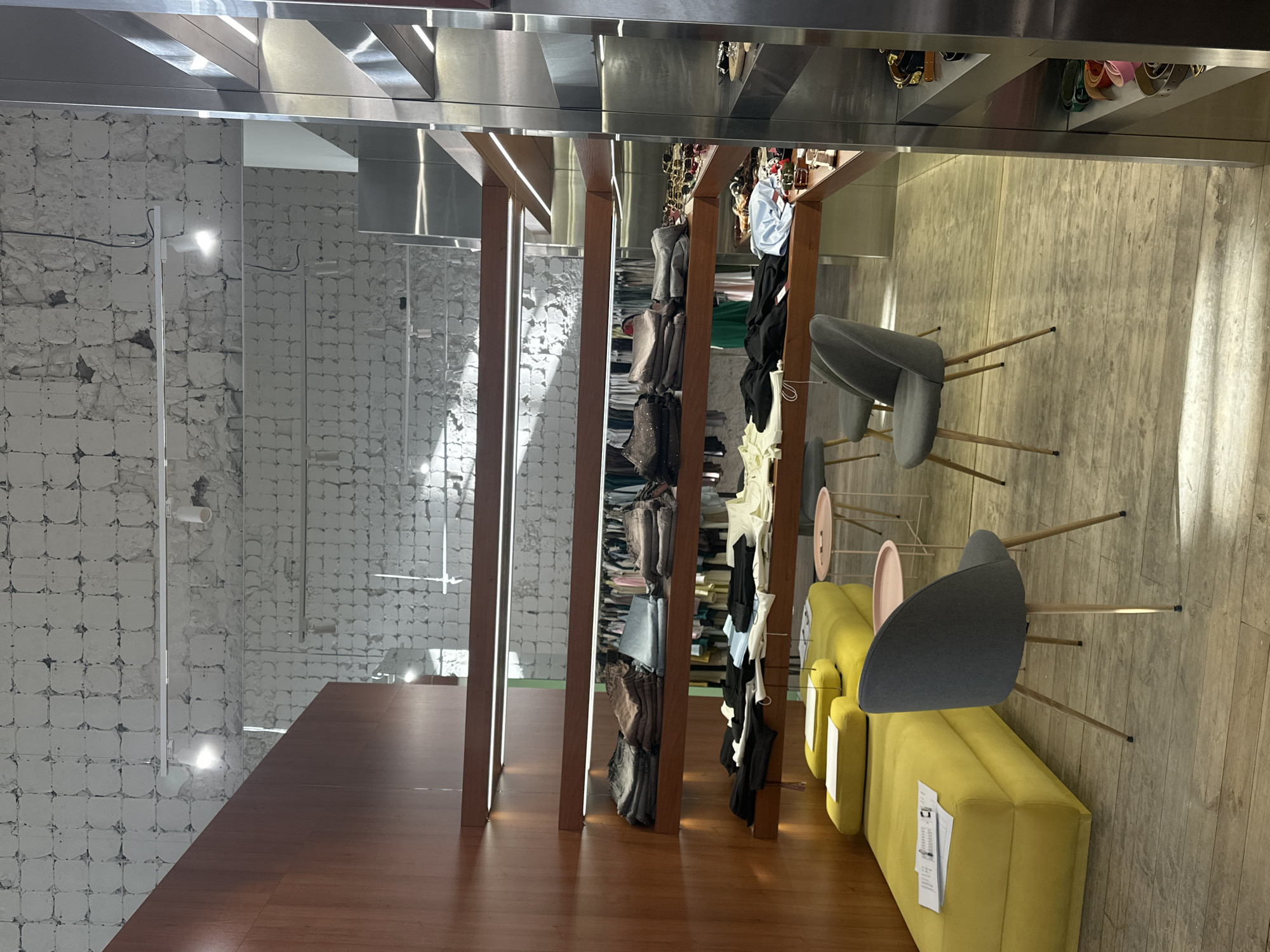 The image depicts an indoor retail space, likely a boutique or clothing store, with a modern and minimalist aesthetic. The scene is dominated by wooden fixtures and shelves showcasing various garments and accessories. On the left, a wall covered in rough, white textured tiles creates a contrasting backdrop. Sunlight streams in from an unseen source, casting sharp shadows and creating bright reflections on the metallic ceiling and surfaces, suggesting it's daytime.
The shelves are organized with folded clothes and what appear to be handbags or shoes displayed on upper shelves. One section features rows of hanging clothing, including jeans and tops. In the foreground, a polished wooden floor reflects the lighting and the overall ambiance. To the right, a bright yellow modular sofa is paired with minimalist gray chairs with slender wooden legs. The chairs are angled towards a display area, suggesting a seating arrangement for customers.
A white tag with printed text is visible on the yellow sofa. The overall impression is one of a clean, stylish, and possibly upscale retail environment. The lack of people suggests the store is either closed or currently unoccupied. The location cue of Odesa, Ukraine, does not provide specific visual markers in this particular image.