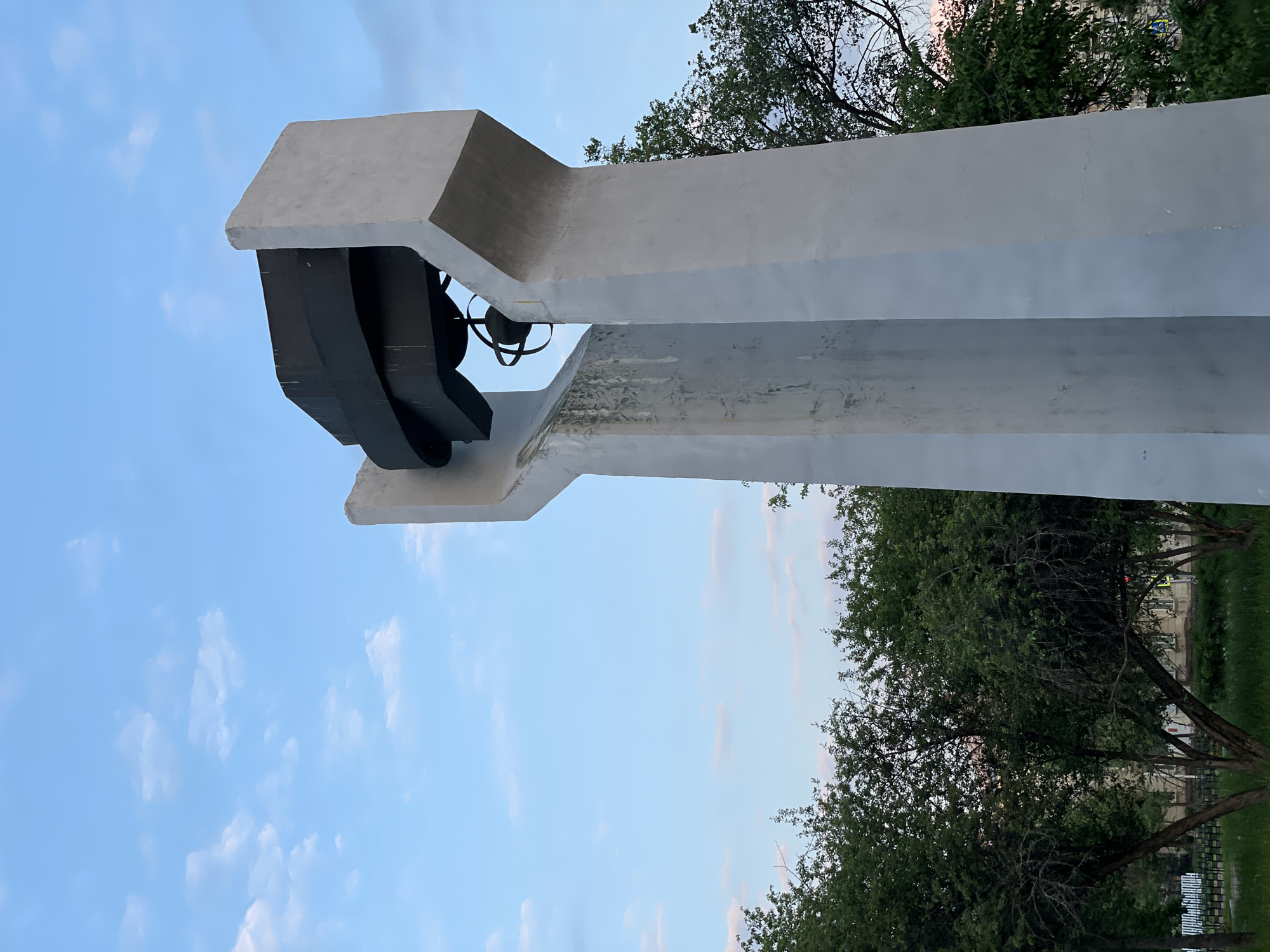 The image shows a monument featuring a large bell suspended within a concrete structure, likely a bell tower or memorial. The monument is located in Zheleznogorsk, Russia. The structure is predominantly white concrete, showing some signs of weathering, with stains visible on the left side. Above the bell, a dark, angular canopy shelters it. In the background, lush green trees with dense foliage are visible, suggesting a park or a well-maintained urban green space. The sky above is a pale blue with scattered, wispy clouds, indicating it might be early morning or late afternoon. The overall scene is tranquil and appears to be an outdoor public space.
