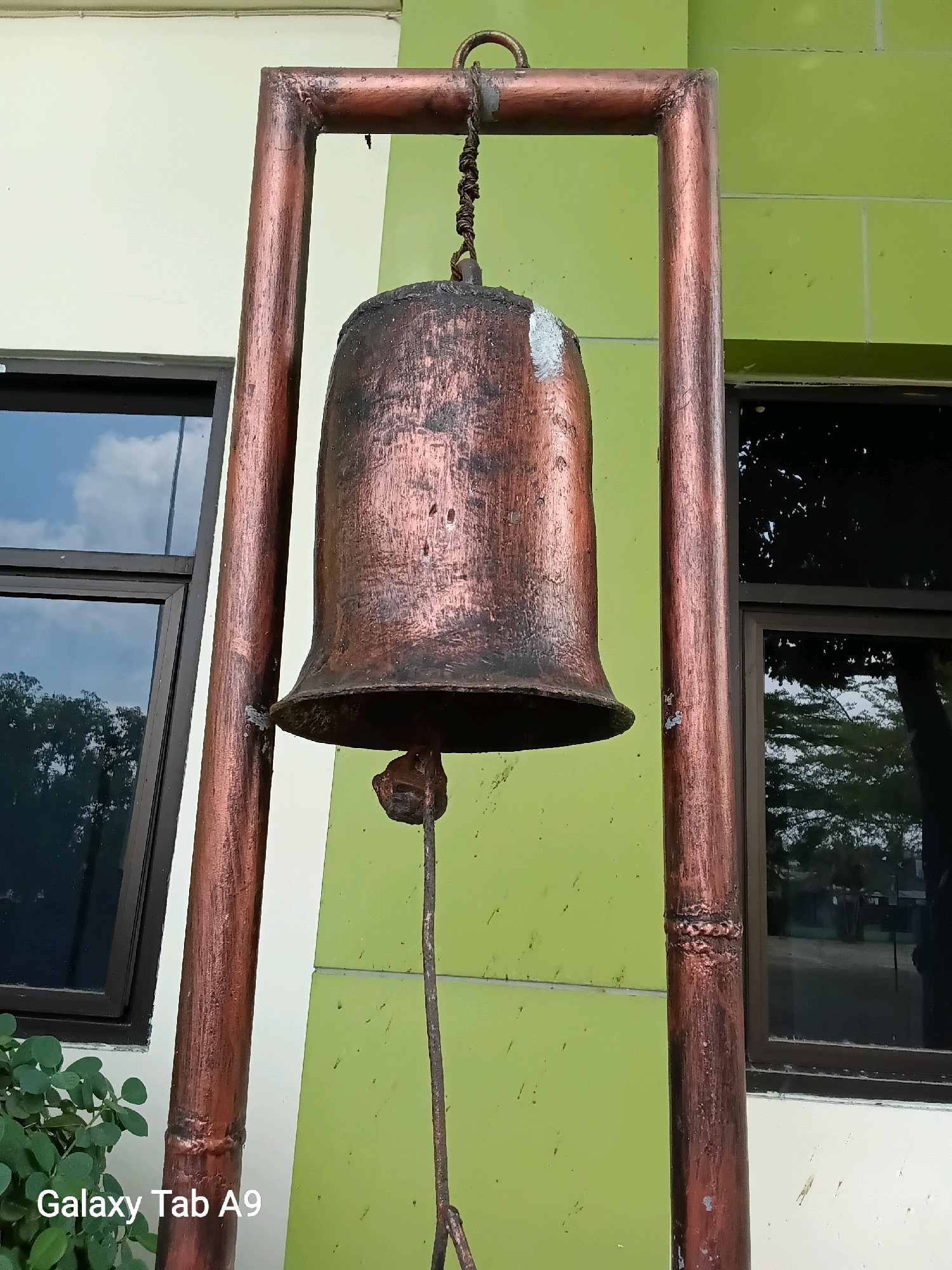 A weathered, copper-colored bell hangs from a rustic, metallic frame against a pale green building with white trim and dark-framed windows reflecting the sky and trees. The bell is suspended by a dark, twisted rope. A slender metal clapper hangs beneath the bell. The building appears to be located in Karangan, Indonesia, based on the metadata. To the left, a bush with green leaves is partially visible. The overall impression is one of aged construction and a tropical environment.