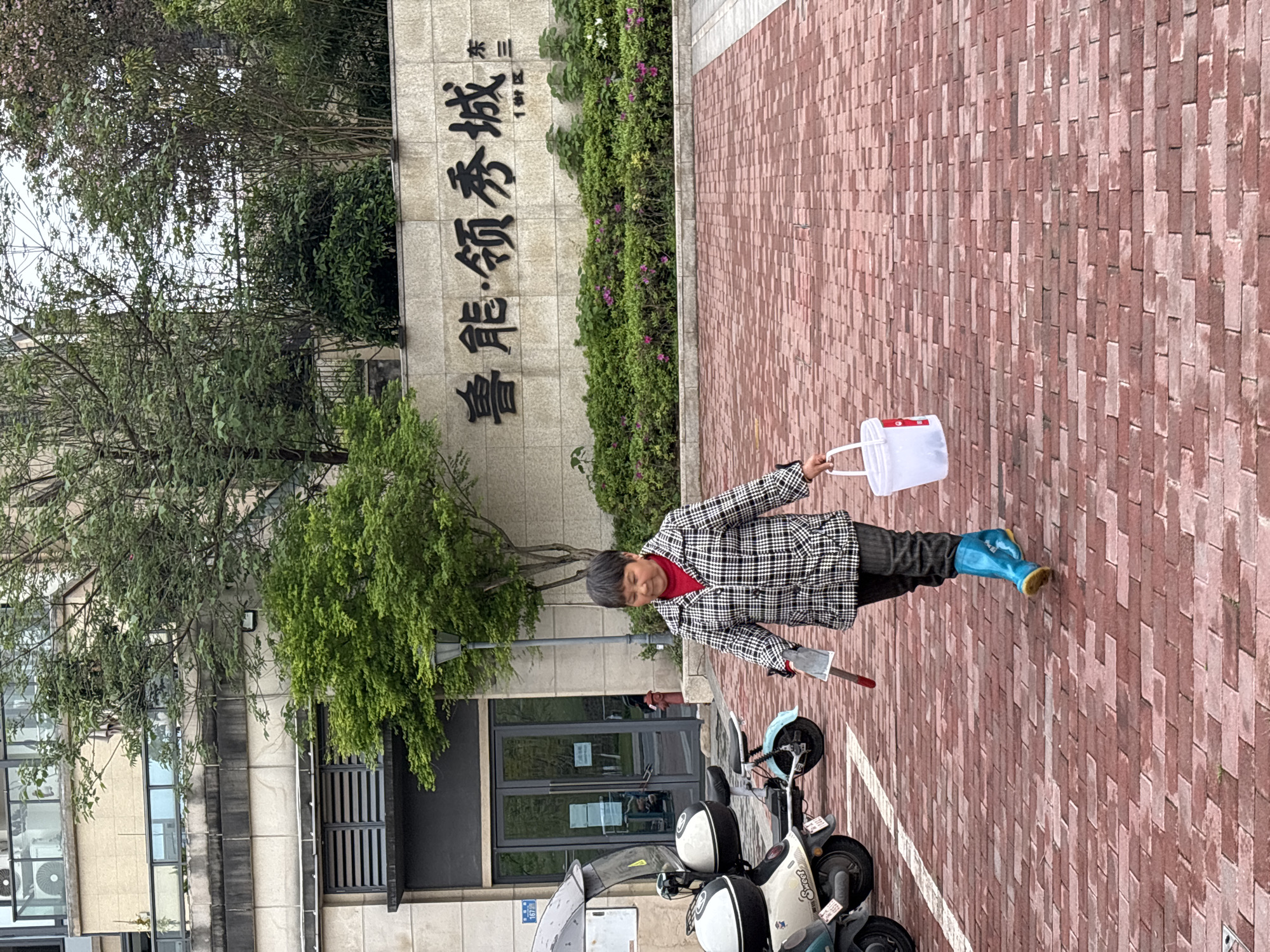 A female subject, appearing middle-aged, walks left to right across a damp red brick paved area in the Lǔnéng Lǐngxiù Chéng (鲁能•领秀城), 1st Block, East Three residential complex, Jiangjin, Chongqing, China. She wears a red shirt, a black and white plaid jacket, dark trousers, and bright blue rubber boots with yellow soles. In her right hand, she holds a small metal scraper with a red handle, and in her left, a white plastic bucket with a red label. Two electric scooters are parked behind her to the left; the closer one is light-colored with "Sweet" branding. A multi-story building with light-colored stone cladding and a dark-framed glass double-door entrance is visible in the background, partially obscured by green trees. A low wall with embossed Chinese characters (Lǔnéng Lǐngxiù Chéng) and additional text ("1街区" and "东三") fronts a landscaped bed of green shrubs and pink-flowering plants. The sky is overcast.