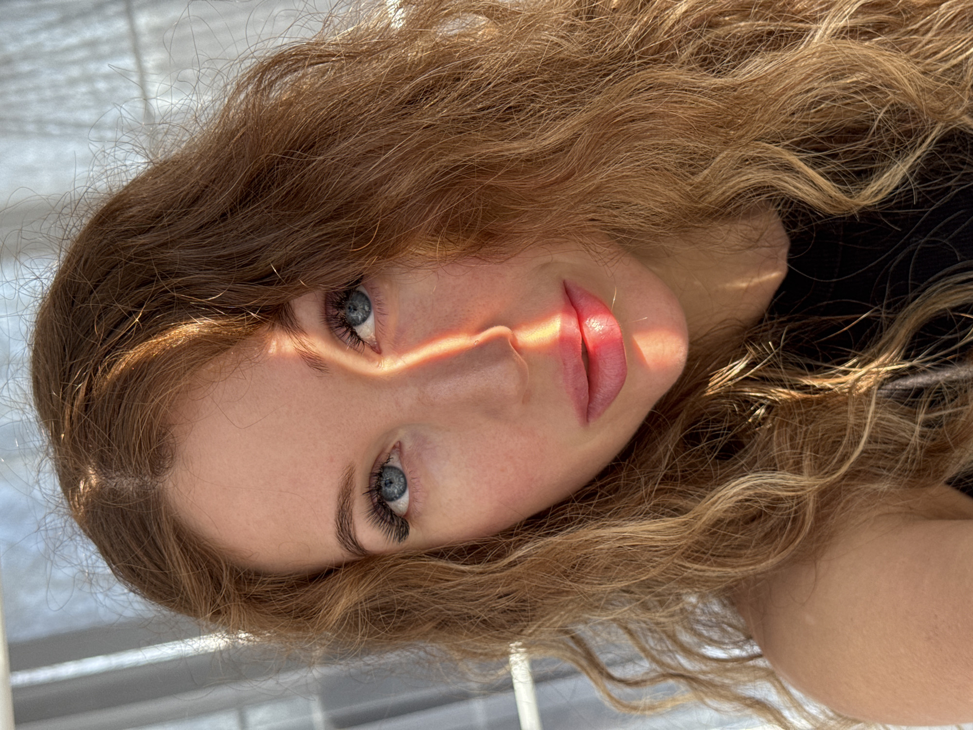 A close-up portrait, taken in Chaikovsky, Russia, features a young woman with long, wavy, reddish-brown hair. Her head is tilted, and her blue eyes are directed towards the viewer with a calm expression. A strong, warm light source, likely from a window, casts a distinct band of light and shadow across her face, illuminating her nose and full, pink lips. This lighting emphasizes her skin texture, long eyelashes, and neatly shaped eyebrows. Her voluminous, wavy hair, with lighter highlights, frames her face and flows down her shoulders, some strands catching the direct sunlight. She appears to be posing for the photograph. The background is soft and out of focus, showing subtle horizontal lines that suggest an indoor setting, possibly near window blinds. No text is visible in the image.