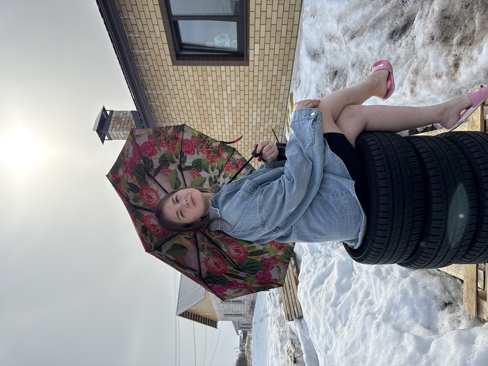 An outdoor scene in Gornyi (Dvurechenskoe S/P), Russia, features a young adult female seated on a stack of three black vehicle tires, which rest on a wooden pallet amidst melting snow and bare ground. The female, facing slightly right of center and looking towards the camera, wears a light blue denim jacket, dark shorts, and pink slip-on sandals. She holds an open umbrella adorned with a pink rose and green foliage pattern. Behind her to the right stands a light beige brick building with a dark roof, a visible brick chimney, and a dark-framed window containing a faint reflection of a child. Icicle-like decorative lights hang from the building's eave. To the left, another building, possibly under construction with white block walls and a corrugated metal roof, is partially visible in the distance, alongside utility poles and power lines. The sky is bright and overcast, with the sun visible near the top center of the frame.