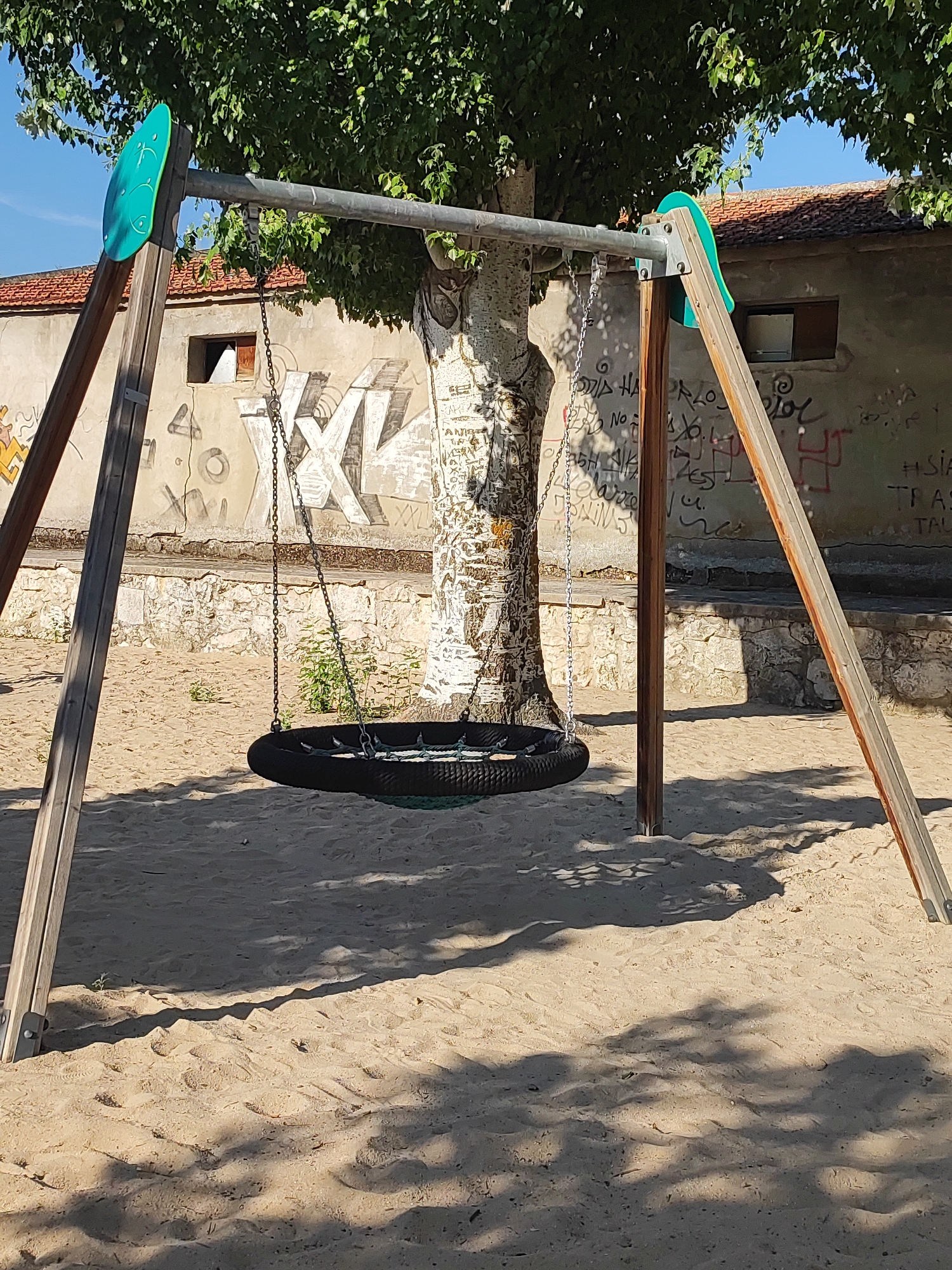 A tire swing hangs from a metal bar supported by two A-frame structures made of wood. The swing is suspended by chains. The scene is set in a sandy playground area with a tree in the background. Behind the tree, a building with a tiled roof and graffiti on its walls is visible. The building has at least two windows, one of which is partially visible. The ground is covered in sand, with shadows cast by the tree and the swing structure. The sky is clear and blue. The location is the playground in Traspinedo, Spain.
