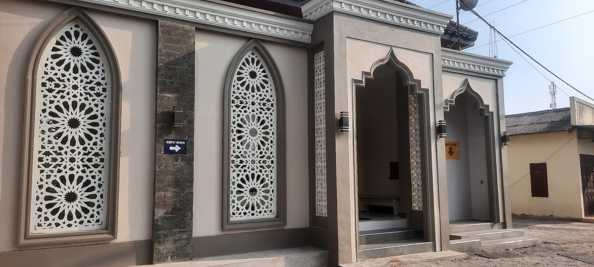 The image shows the exterior of a building with Islamic architectural elements, possibly a mosque or a related religious facility, located in Tumpang, Indonesia. The building has a pale beige facade with gray stone accents and decorative white latticework on arched window openings. The windows feature intricate geometric patterns common in Islamic art. An entrance archway is visible, with steps leading up to it, and two modern black outdoor light fixtures are mounted on either side of the arch. A blue sign with white text and an arrow pointing to the right reads "TEMPAT WUDHU," which translates to "Washing Place" in Indonesian, indicating a place for ritual ablution. To the right of the entrance, a yellow sign with red text and a downward-pointing arrow is partially visible, possibly indicating another direction or instruction. In the background, a lighter-colored building with a dark-tiled roof is situated. The sky is clear and bright, suggesting daytime. The ground in front of the building is paved with a rough, gravel-like surface. The overall scene depicts a well-maintained religious building in a residential or semi-urban area.