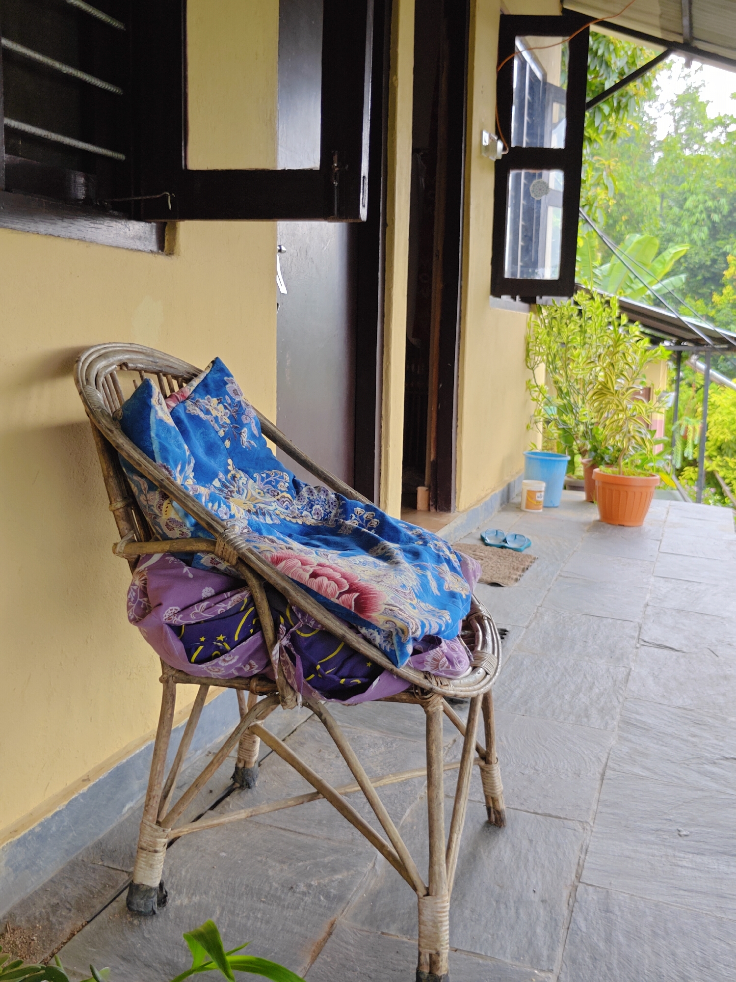 A woven wicker chair is positioned on a stone tiled patio in front of a light yellow wall. The chair is draped with two fabric items: a vibrant blue cloth with floral and intricate patterns, and underneath it, a purple cloth with a pattern of yellow stars. To the right of the chair, a small outdoor area is visible, featuring two potted plants, one in an orange pot and another in a blue pot, placed on the tiled ground. Further back, a dark wooden door and a partially open window are visible. The scene suggests an outdoor space connected to a building, possibly a residence or guesthouse, with lush green foliage visible through the window and in the background, indicating a tropical or subtropical environment. The overall atmosphere is one of stillness and quiet, with no people present. This scene is set in Pokhara, Nepal, a city known for its natural beauty and mountainous backdrop, though the immediate surroundings here focus on a more domestic outdoor setting.