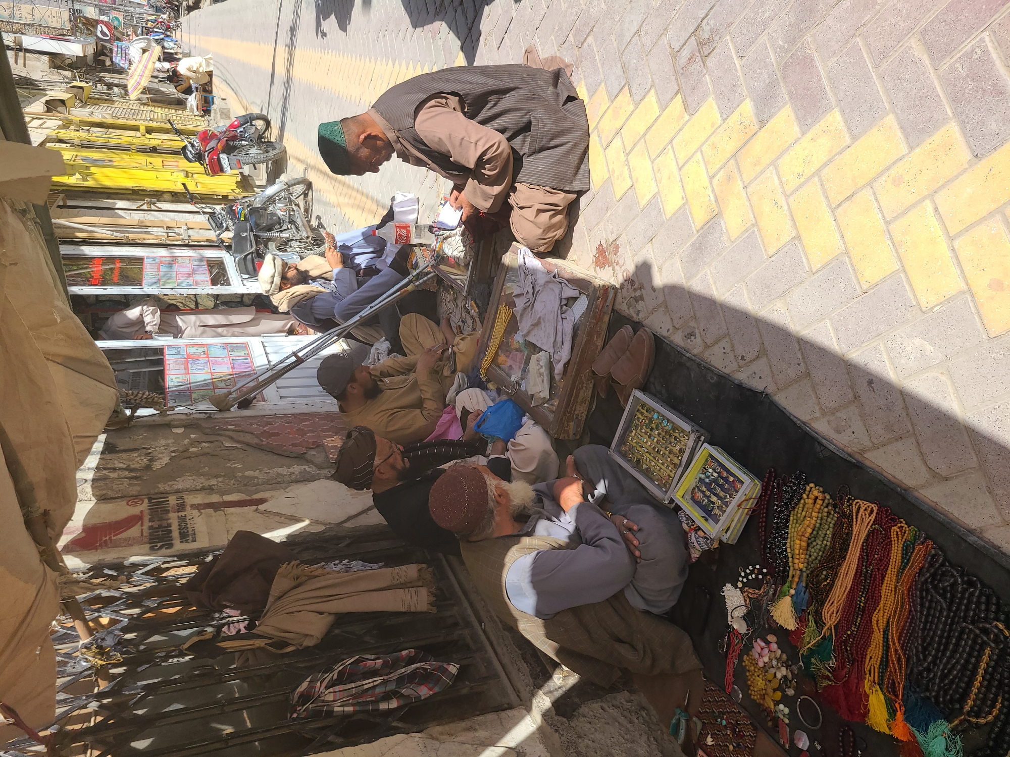 This photograph, likely taken in Quetta City Tehsil, Pakistan, shows a row of men sitting and kneeling on a sidewalk adjacent to a storefront. The men appear to be vendors, possibly selling goods displayed on the ground in front of them. One man is leaning over, seemingly handling items or money. The sidewalk is paved with brick, and there is activity visible in the background. There are motorcycles and goods on shelves nearby. There is a sign that says "SIDEPOINT".