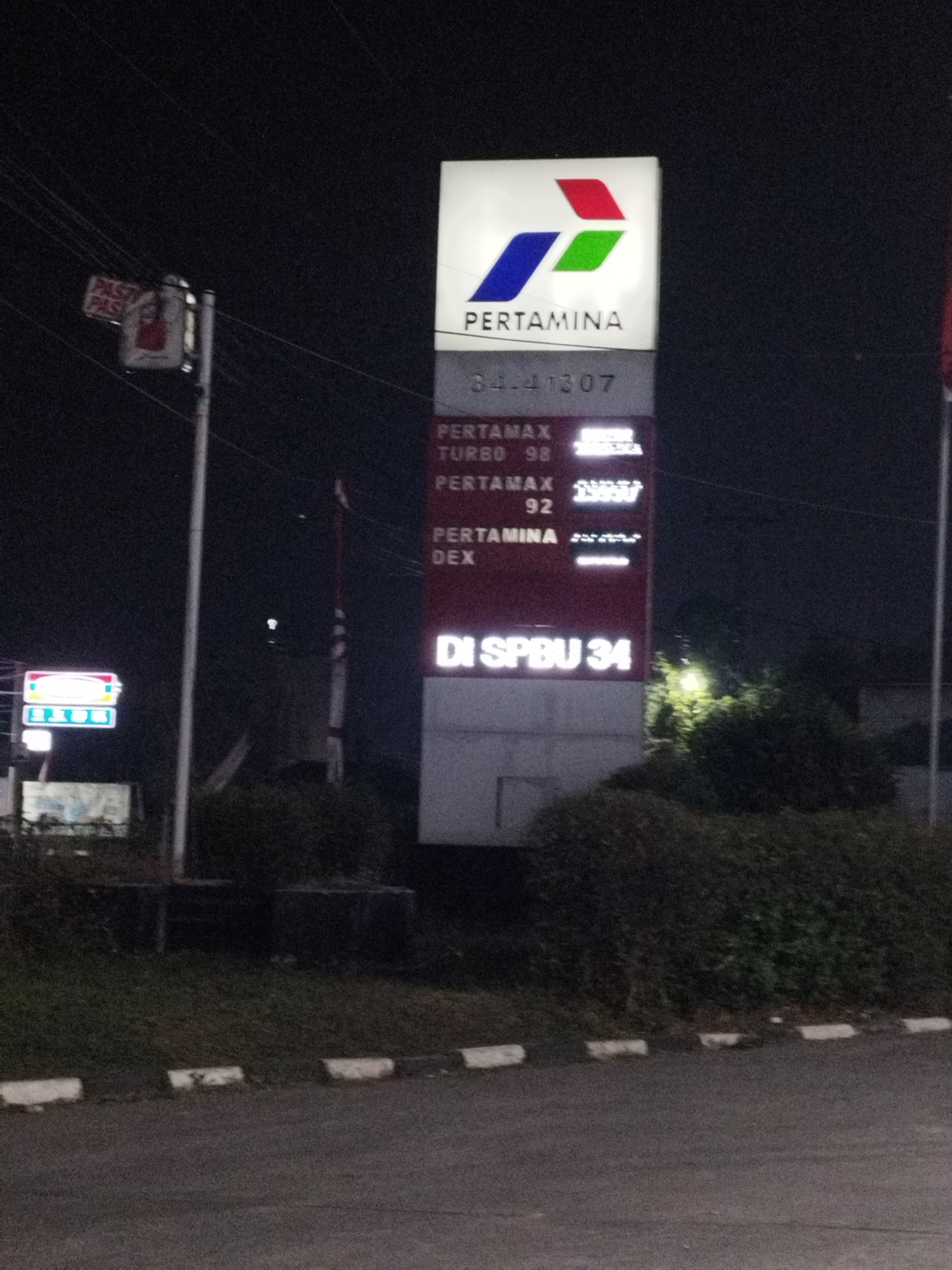A night-time outdoor shot shows a Pertamina gas station sign in Karawang, Indonesia. The sign is illuminated and displays the Pertamina logo and fuel prices. In the foreground, a street with white curbs is visible. Behind the sign, there is a large bush and a street lamp. To the left of the Pertamina sign, there's a smaller sign for "Indomaret" with illuminated logos. The overall scene depicts a typical street view of a gas station and convenience store at night.