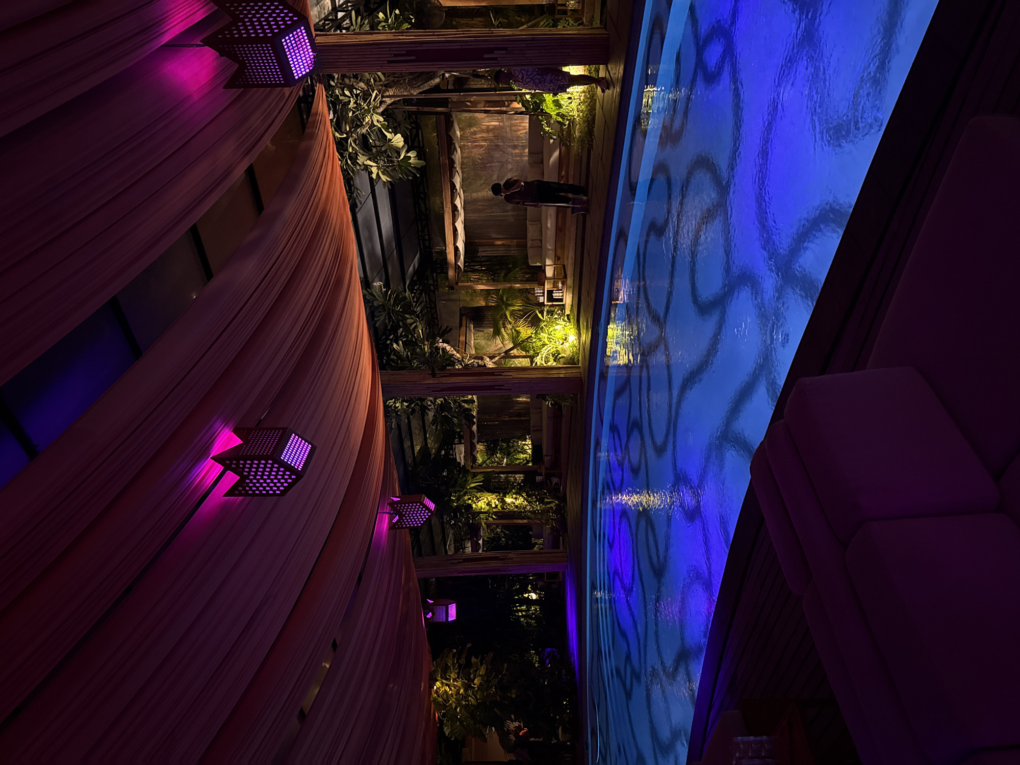 This media file captures a vibrant, dimly lit resort or club setting at night, likely in North Kuta, Indonesia. The scene is presented from a slightly elevated perspective looking down into the area.
On the left side of the frame, soft, flowing drapes in a warm, earthy tone (possibly beige or light orange) hang gracefully. Several decorative, square lanterns with intricate perforations are affixed to the structure above the drapes, casting a striking magenta and purple uplight onto the fabric, creating a rich, atmospheric glow.
In the mid-ground, a walkway or lounge area is visible, bordered by lush tropical foliage and wooden architectural elements, possibly trellises or supports. This area is lit with soft, warm yellow and green accent lights that highlight the greenery. Two individuals can be seen in this section. One person, wearing lighter clothing, stands closer to the pool's edge, while another, in darker attire, is further back, near some dimly lit seating and a small, glowing rectangular object on a stand.
Dominating the right side of the image is a swimming pool filled with vividly blue water. The pool's bottom features a repeating pattern of swirling, organic shapes in a darker hue. The water surface is gently rippled, reflecting the patterns and ambient light. A dark wooden deck appears to line the pool's edge.
In the immediate foreground on the far right, a section of upholstered lounge seating in a deep, dark color, possibly purple or maroon, is visible, mostly obscured in shadow.
The overall atmosphere is one of luxury, relaxation, and tropical nighttime allure, accentuated by the strategic and colorful lighting. There is no visible text in the image.