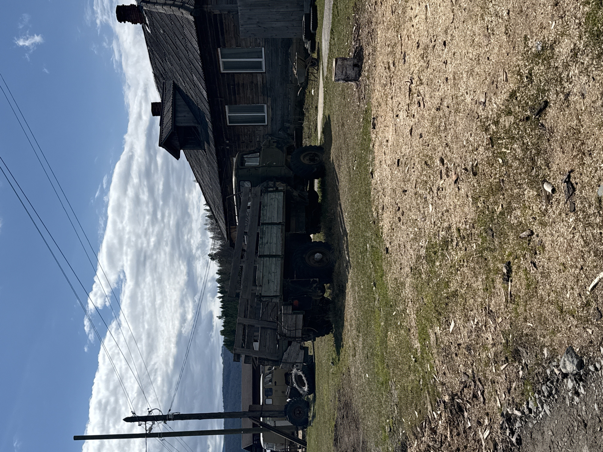 The image depicts a rustic rural scene in Kordonnyi, Russia, under bright daylight with a partly cloudy sky. The main subject is a large, dark-stained wooden house or building with multiple windows, a prominent chimney, and a dormer window, suggestive of traditional log construction.
Parked in front of the building are two older, rugged trucks. The most prominent is a large, military-style utility truck with a green cab and a wooden flatbed, featuring large, off-road tires. Behind it, partially obscured, is another smaller truck or vehicle. The ground in the foreground and around the vehicles is a mix of dry grass and extensive wood chips or sawdust, suggesting a logging or woodworking context. A tree stump is also visible on the ground.
In the left foreground, several utility poles with power lines stretch across the frame. The background features rolling hills or mountains covered with evergreen trees, visible beneath a long, horizontal cloud formation that separates a clear blue sky from a mass of white clouds. No people are visible, and no specific activity is taking place; the scene appears static. The lighting suggests it is late morning or early afternoon, casting distinct shadows. There is no visible text in the image. The overall impression is one of a remote, working rural environment.