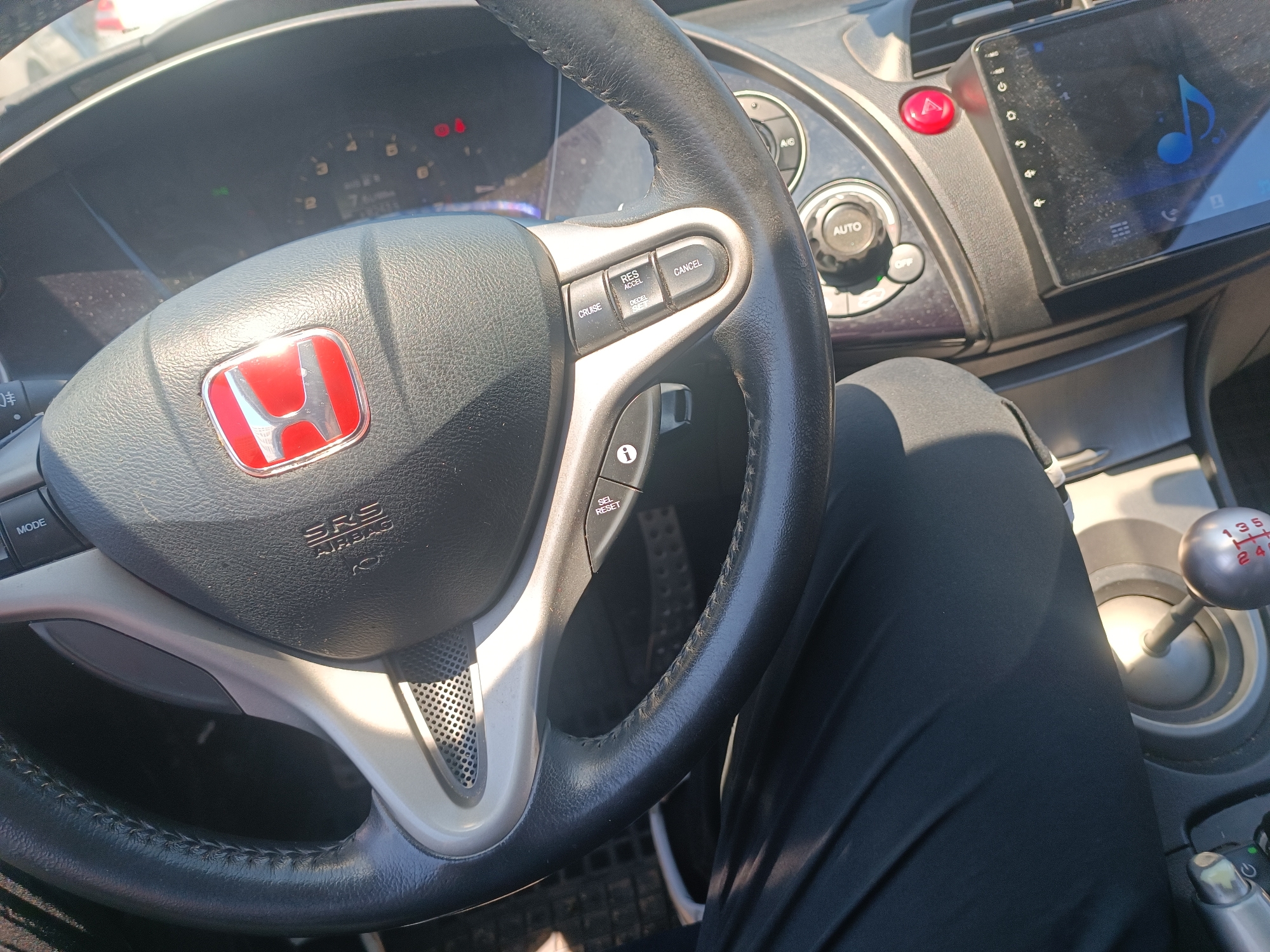 The image shows the interior of a car, specifically the steering wheel, dashboard, and gear shifter. The steering wheel has a red Honda emblem in the center and buttons for cruise control and other functions. The dashboard features an infotainment screen and various controls. A person's leg, covered in black fabric, is visible in the foreground. The gear shifter is a manual type, with visible gear markings. The lighting suggests it is daytime, with sunlight entering the car. There is no visible indication of the car's specific location within Burgas, Bulgaria, nor is there any information to suggest an event or interaction taking place.
