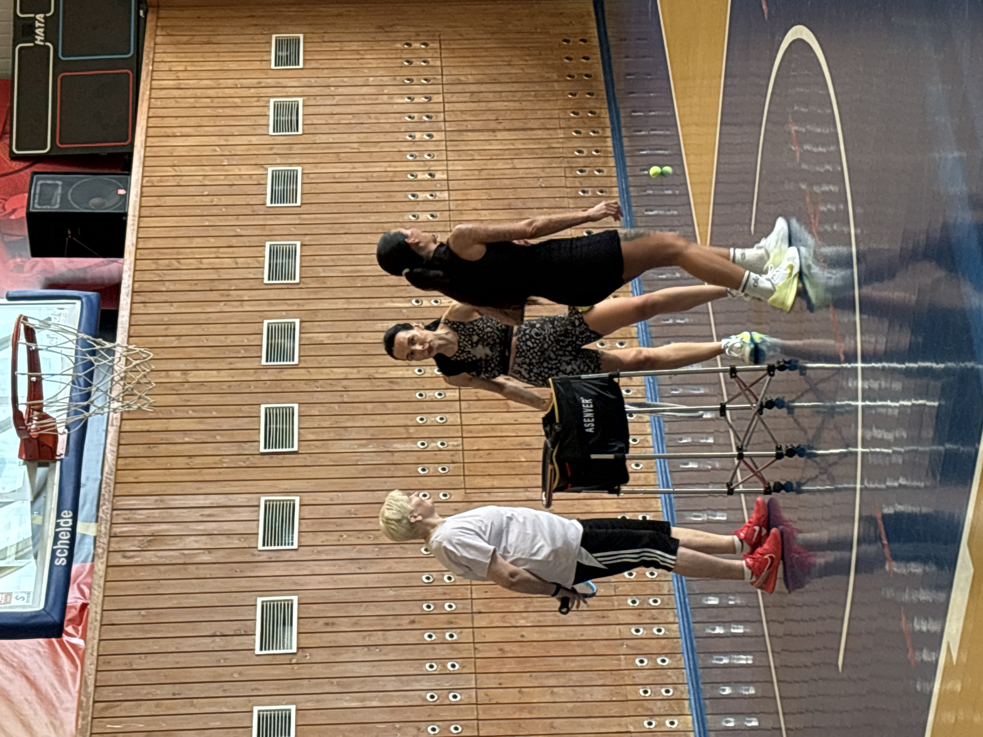 The image depicts an indoor scene, possibly a gymnasium, with three individuals present. The location is Chaikovsky, Russia, based on the "Schelde" logo seen on the basketball backboard.
A young man with blonde hair stands nearest the camera, wearing a white t-shirt and black shorts with white stripes, and red athletic shoes. He is near a portable ball storage container.
Two women are further away. One wears a black dress and white/greenish athletic shoes. The other woman wears a short black and white patterned dress and athletic shoes that are blue, black, and yellow. Both are engaged in exercise.
The background includes a wooden wall with rows of white circular indentations and rectangular vents. To the left, a basketball hoop and backboard are visible along with large black speakers. The floor is shiny and appears to have markings. Two tennis balls are on the floor in the upper right area.