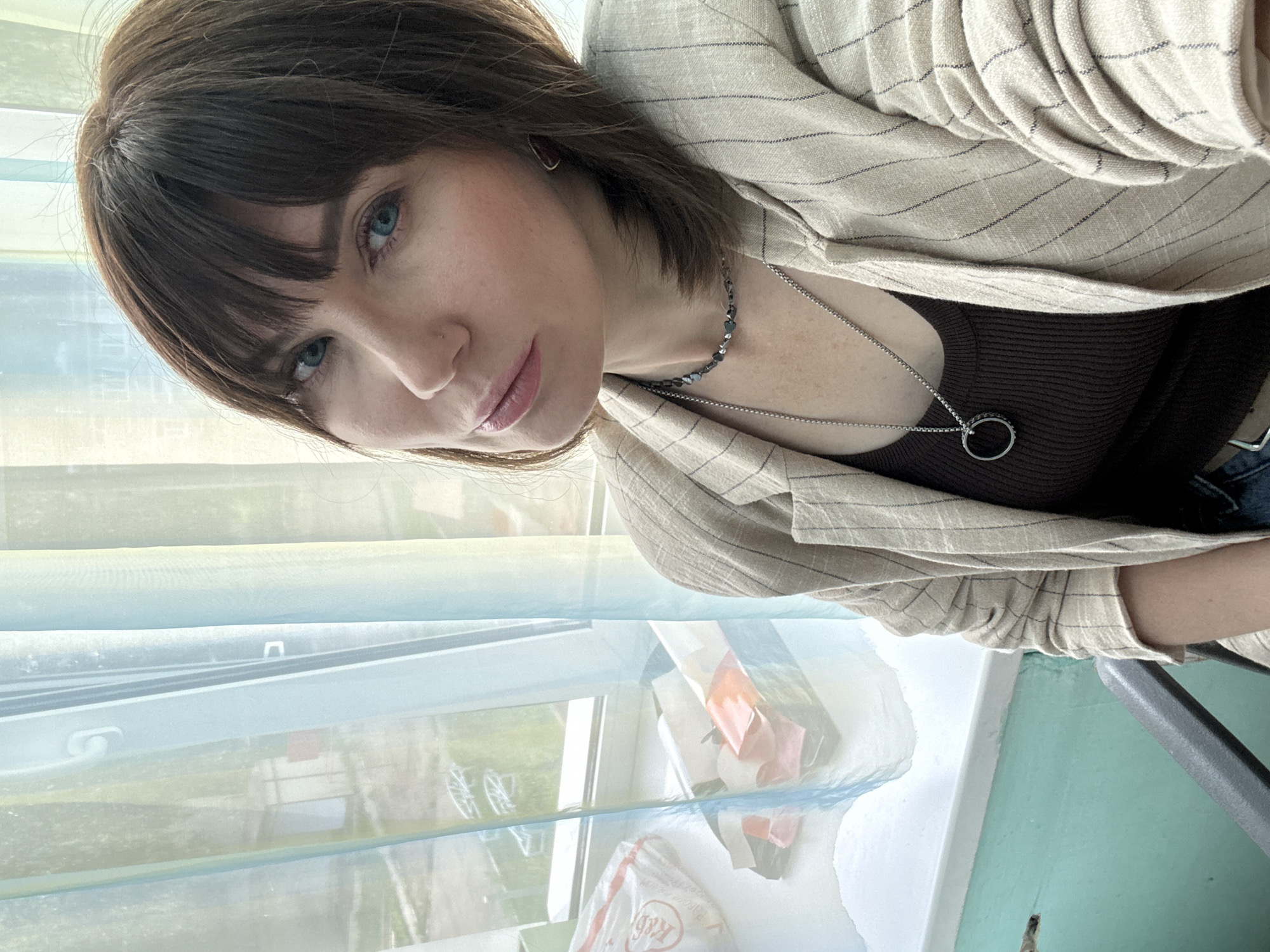 A woman with shoulder-length brown hair and blue eyes, wearing a pinstripe blazer over a brown top, is photographed indoors. She is sitting in front of a window with sheer curtains that diffuse the light from outside. The window sill displays several items, including a white plastic bag with red lettering, and other assorted objects. The room's walls are painted a light teal color. The overall impression is a casual, personal photograph taken in what appears to be a residential or office setting within the city of Chaikovsky, Russia.