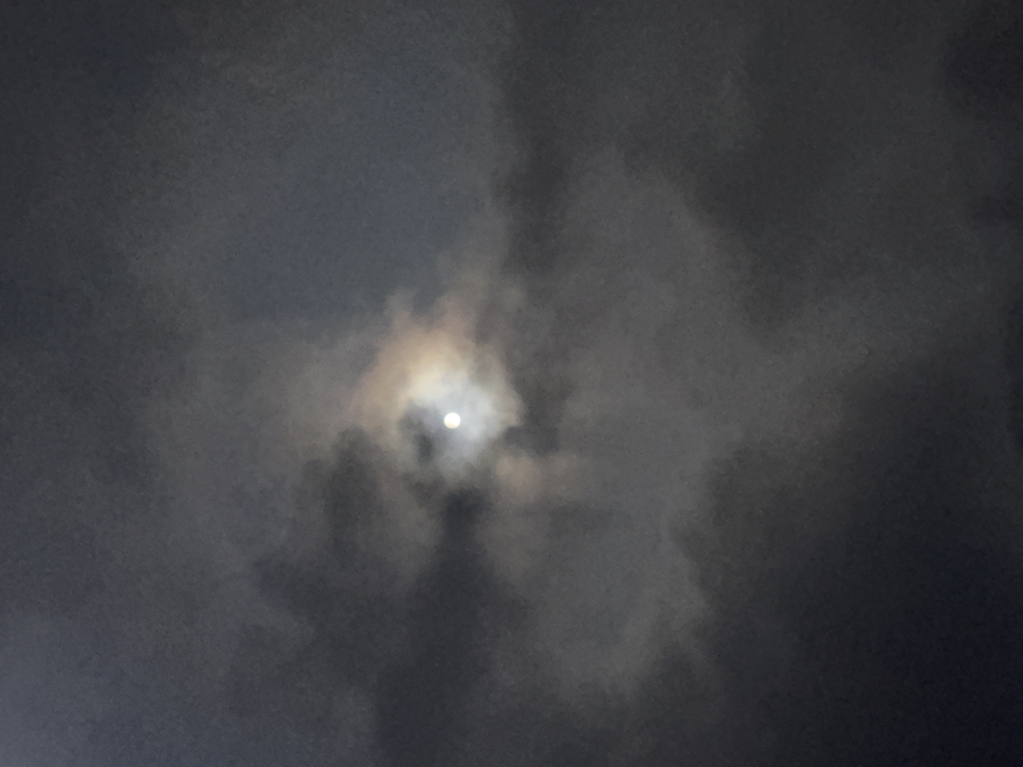A full moon is visible through patchy, dark clouds at night. The clouds are illuminated by the moon, creating a soft glow around it and casting shadows that obscure parts of the lunar disk. The overall atmosphere is moody and ethereal, suggesting a dramatic or mysterious night sky. The image appears to be taken outdoors, looking upwards, possibly from a location in Bhagsu, India, given the context. There are no people or man-made objects clearly depicted in the image.