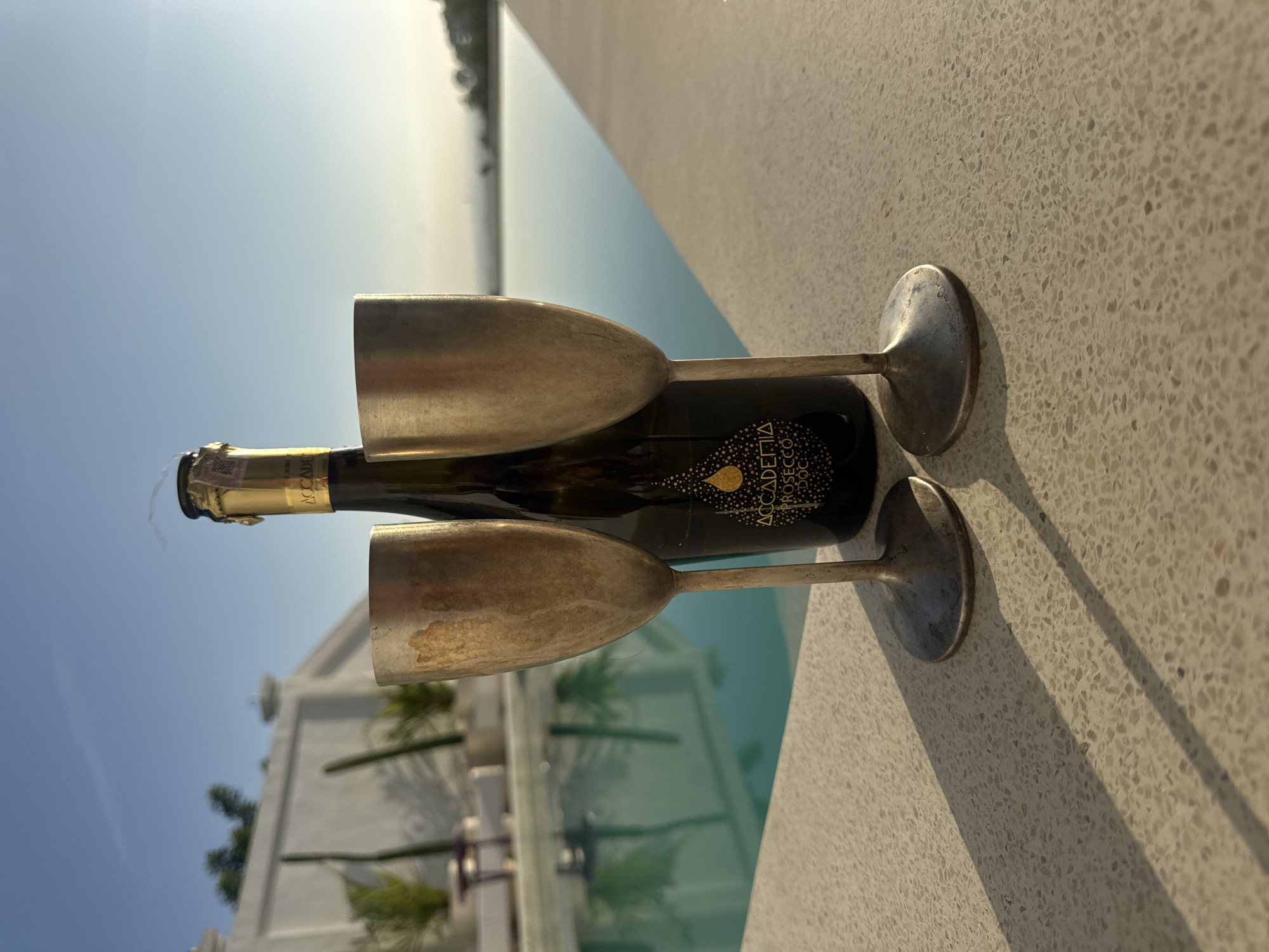 The image shows a bottle of Accademia Prosecco DOC sparkling wine lying horizontally on a light-colored, speckled surface, with two tarnished metal champagne flutes placed on either side of it. The location appears to be outdoors, alongside a pool of water, with blurred white buildings and trees visible in the background, suggesting a resort or villa setting. In the distance, there is a horizon line, indicating proximity to the sea. Based on the location of the city of Kuta Selatan, Indonesia, in the prompt, the landscape is likely a part of that area. There are no people visible in the image. The dominant objects are the wine bottle and the champagne flutes. The scene suggests a moment of relaxation and celebration.