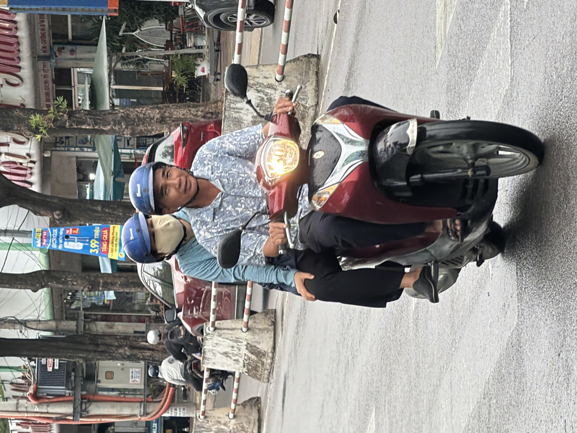 A man and woman are riding a red motorcycle on a wet street in Nha Trang, Vietnam. The man is driving and wearing a blue helmet and a patterned shirt. The woman is behind him, wearing a blue long-sleeved shirt and a light-colored mask. Both are wearing helmets. The street is wet, suggesting it has recently rained. In the background, there are trees, buildings, and other vehicles, including a red car. A utility pole with a blue sign is visible on the right side of the image. Concrete barriers with red and white stripes are placed along the side of the road. The lighting suggests it is daytime.