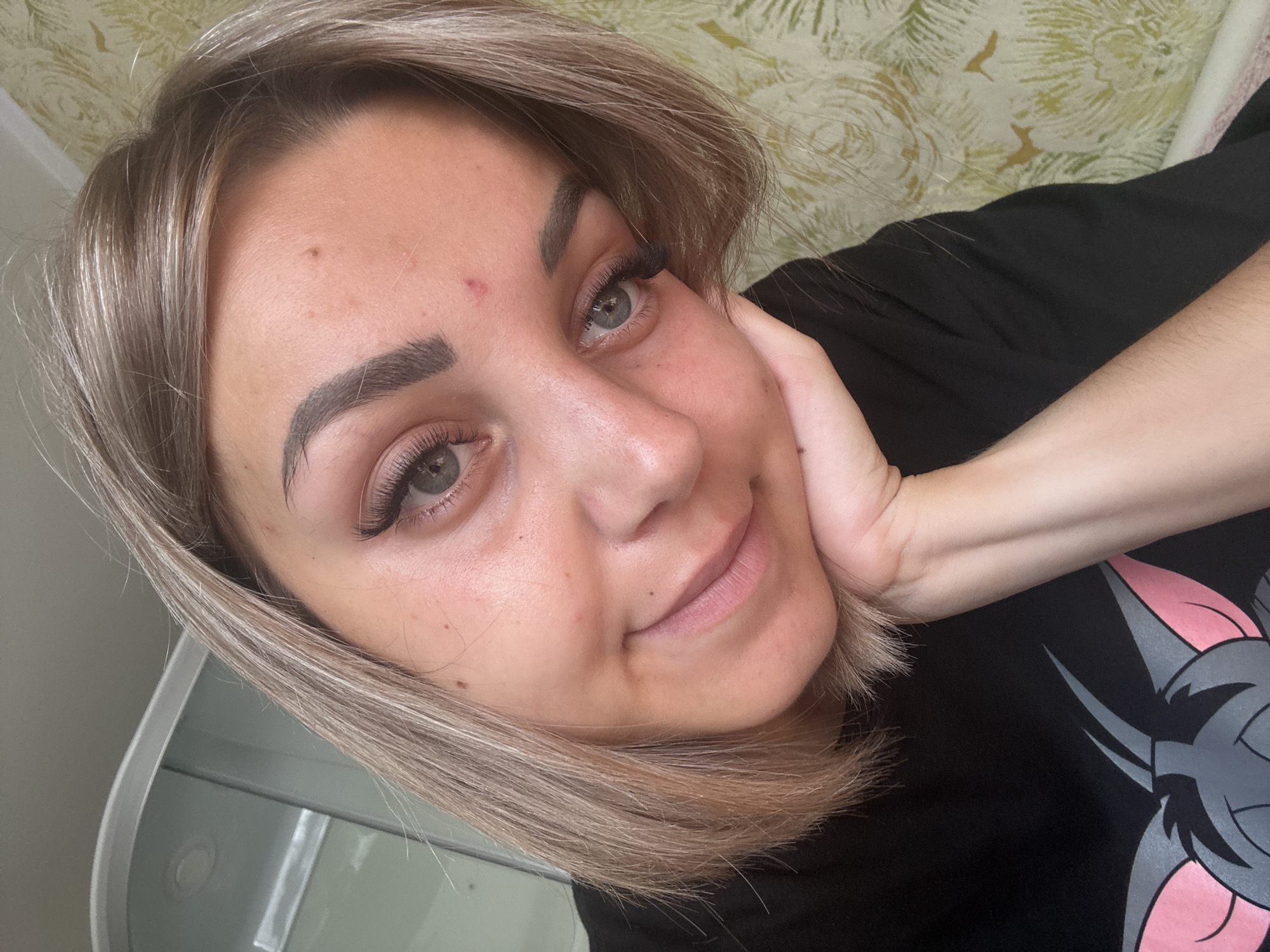 The image is a close-up, self-taken photograph of a young woman with blonde, shoulder-length hair, resting her head on her hand. She is wearing a black t-shirt with a cartoon character print on it. Her eyes are open, and she has a slight smile. Her eyebrows are well-defined, and she appears to be wearing makeup, including false eyelashes. There is a small red blemish on her forehead. The background consists of a textured, light-colored wall with a floral pattern. The overall lighting suggests an indoor setting, possibly a room in a residential building. The specific location in Krasnogorskoye, Russia cannot be determined from the visual information provided in the image.