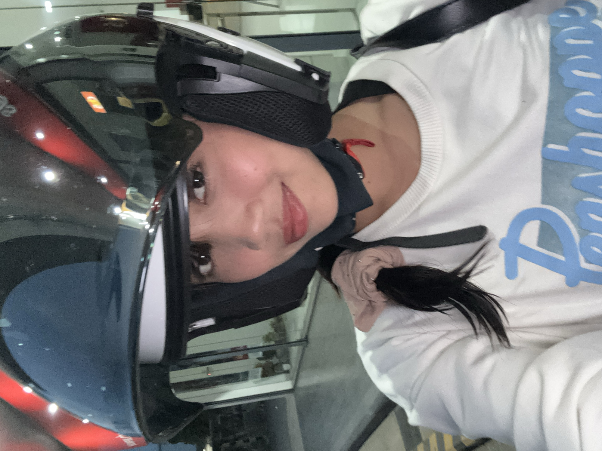 A young woman with long, dark hair pulled back in a ponytail, wearing a white sweatshirt and a black and white helmet with a tinted visor, is seen in a selfie taken from a low angle. She is looking towards the camera with a slight smile. Her hair is tied with a scrunchie, and a small red string or ribbon is visible near her neck, possibly attached to the helmet strap. The background appears to be an indoor setting with a reflective surface, possibly a glass-fronted building or store, with some lighting and blurred figures visible. The overall lighting suggests it is either evening or a dimly lit interior. The logo on her sweatshirt is partially visible, featuring blue lettering. The setting is consistent with an establishment in Davao City, Philippines, possibly a commercial area or a public transport terminal.
