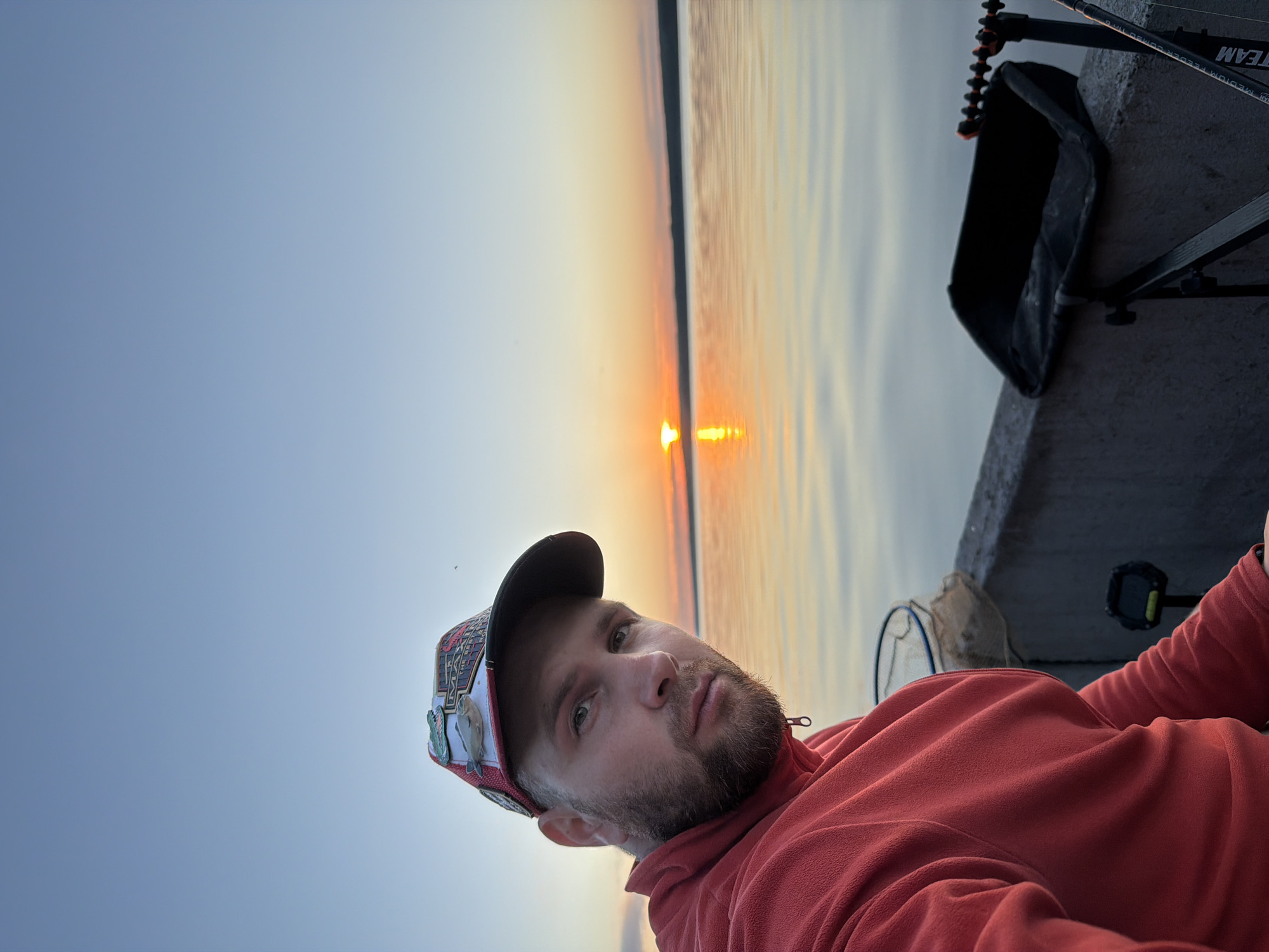 A man wearing a red fleece jacket and a cap with various patches, including a fish, is pictured at sunset on a body of water. The sun is low on the horizon, casting a warm glow on the water and sky. In the foreground, fishing equipment is visible, including a black bait tray and fishing rods. The scene appears to be from the city of Chaikovsky, Russia, given the context. The man is looking directly at the camera.