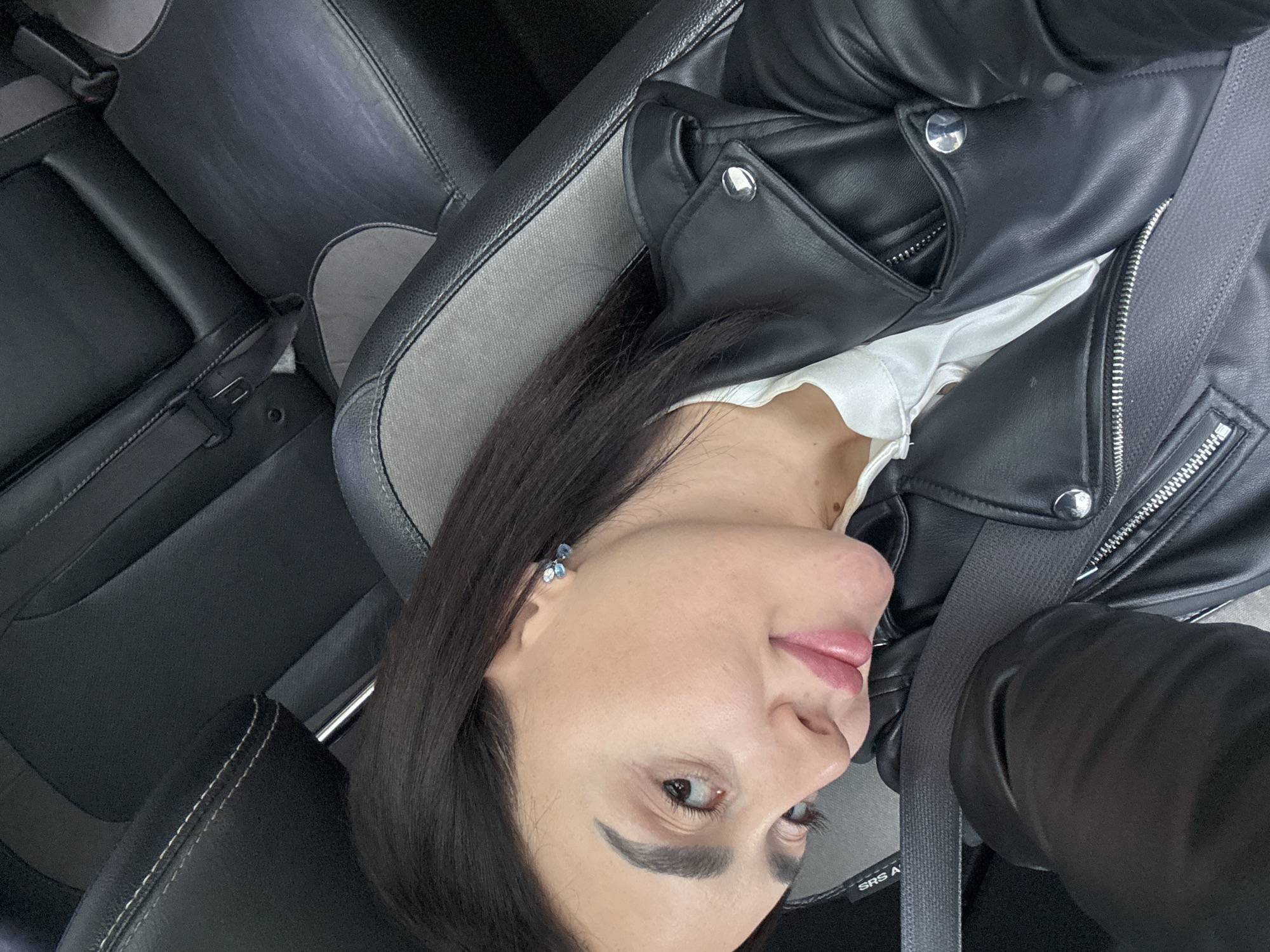 A person is seated in a car, wearing a black leather jacket over a white top and a seatbelt. The car has dark gray or black interior, including leather seats. The location is not specified.