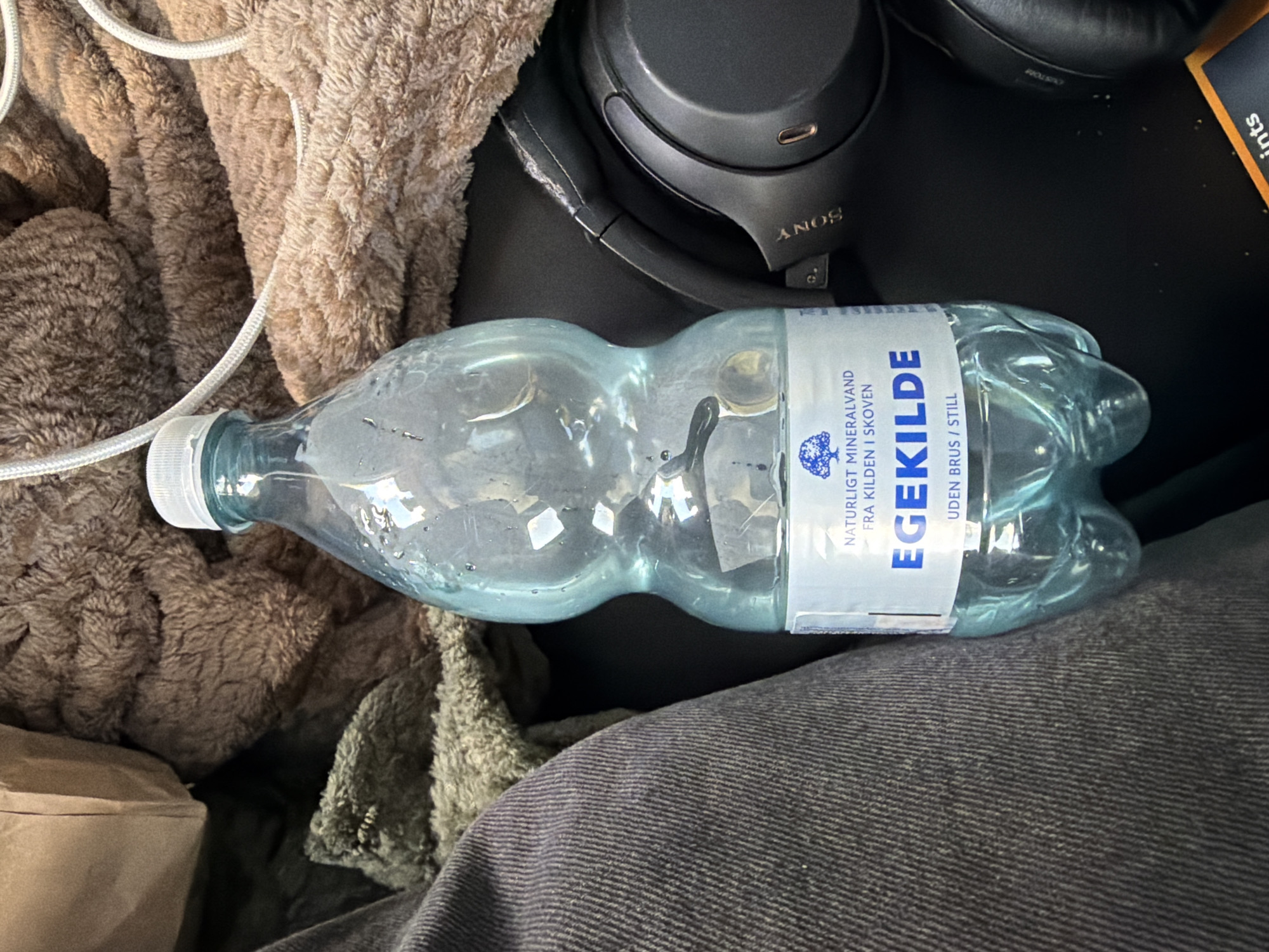 The image shows a close-up of a clear plastic bottle of "EGEKILDE" mineral water. The bottle has a blue tint and is partially filled with water. A white label on the bottle reads "NATURLIGT MINERALVAND FRA KILDEN I SKOVEN" and "EGEKILDE" in large blue letters, followed by "UDEN BRUS / STILL". The bottle is resting on a dark surface, possibly a table or floor, next to a pair of black Sony headphones. In the background, parts of brown and green fuzzy fabric are visible, along with a brown paper bag. The scene is dimly lit. The location of Ödsmål, Sweden is not discernible from the image itself.