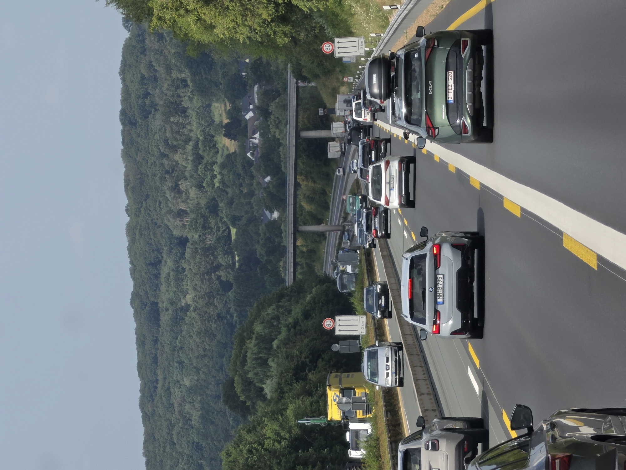 The image captures a congested highway scene, likely on a section of the A3 Autobahn near Königswinter, Germany, judging by the road markings and signage typical of German autobahns, and the general landscape of rolling hills and dense forest characteristic of the Siebengebirge region. The highway stretches ahead, with multiple lanes filled with various vehicles including passenger cars, SUVs, and trucks, indicating heavy traffic. A yellow truck is visible in the left lane, partially obscured by traffic. Overhead, a bridge spans the highway, further suggesting an autobahn environment. Traffic signs are present, including a speed limit sign indicating 80 km/h. The surrounding environment is dominated by lush green trees and hills, with some houses visible on a distant hillside, indicating the highway passes through a semi-rural or suburban area. The sky is overcast with a light blue hue. The perspective is from within a vehicle, looking forward, capturing the density of traffic and the surrounding natural landscape.