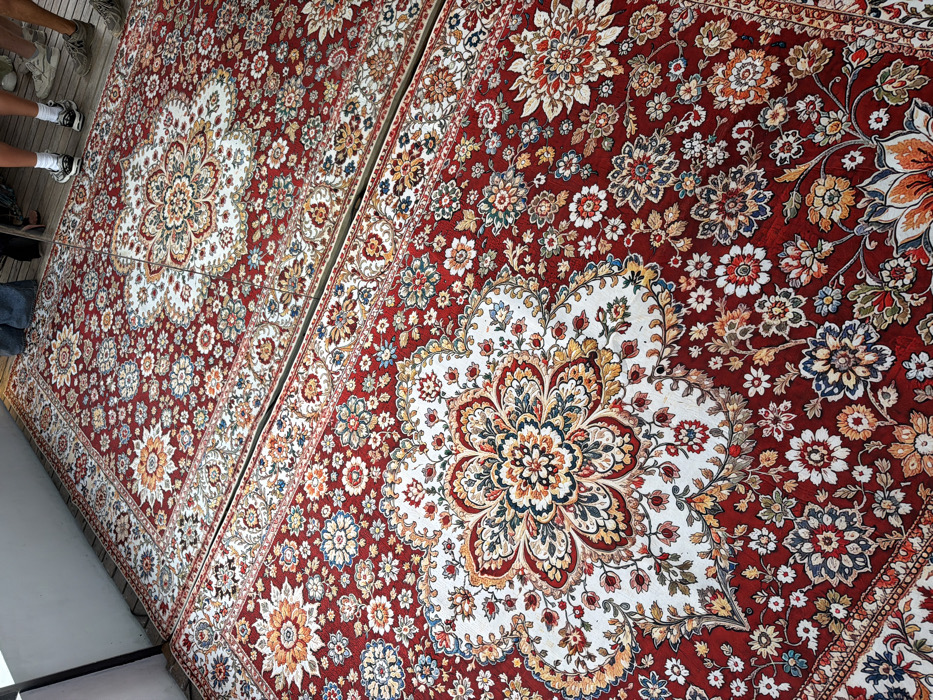 The scene displays a floor area covered by at least two contiguous, large, ornate rugs, characterized by a predominantly red background adorned with intricate floral and curvilinear patterns in white, gold, blue, green, and orange. Each rug features a prominent central medallion with elaborate floral designs on a white field. A distinct horizontal seam or gap separates the upper and lower sections of the rugs, suggesting multiple pieces laid out. The rugs rest on a light-colored wooden planked surface. In the upper right portion of the image, the lower legs and feet of multiple individuals are visible standing on or near the rugs. One pair of feet wears white socks and black athletic shoes, another pair wears light-colored athletic shoes, and a leg partially visible on the far left wears blue jeans. A vertical white architectural element is visible along the left edge of the frame. This setting is located in Canggu, Indonesia.