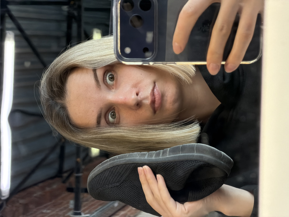 A woman with short blonde hair takes a selfie in a mirror. She holds a black sneaker up towards the camera. The reflection shows metal scaffolding and a bright white light in the background. The woman's face is slightly tilted, she gazes into the camera with light eyes and has fair skin. She holds up a phone in one hand to take the selfie. The phone has a clear protective case, revealing its dark blue back.
