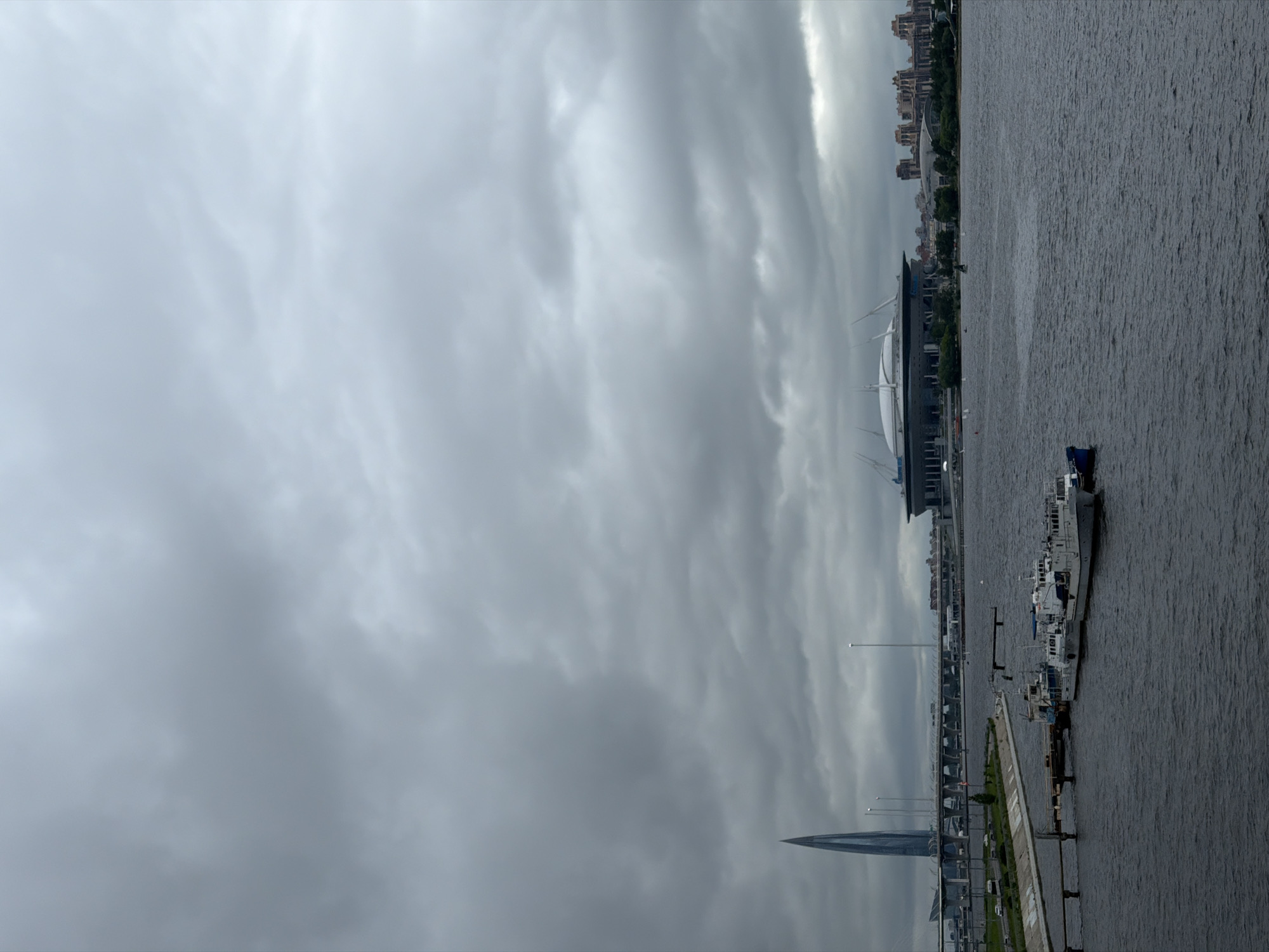 This image captures a panoramic view of the Neva River in Saint Petersburg, Russia, on a cloudy day. In the foreground, the dark, choppy water of the river is visible, with several boats docked at a pier on the left side. These boats appear to be of moderate size and are predominantly white.
In the middle ground, a concrete embankment lines the river, with a road and some greenery. Further back, a prominent, distinctive skyscraper, identified as the Lakhta Center, stands tall on the left side of the frame, its glass facade reflecting the overcast sky. A large, modern stadium, likely the Gazprom Arena, is visible on the right, characterized by its distinctive white, domed roof and surrounding infrastructure. In the distance, a cluster of buildings with a reddish-brown hue forms part of the city's skyline. The sky is filled with heavy, grey clouds, suggesting overcast weather or the possibility of rain. The overall atmosphere is somewhat muted due to the atmospheric conditions.