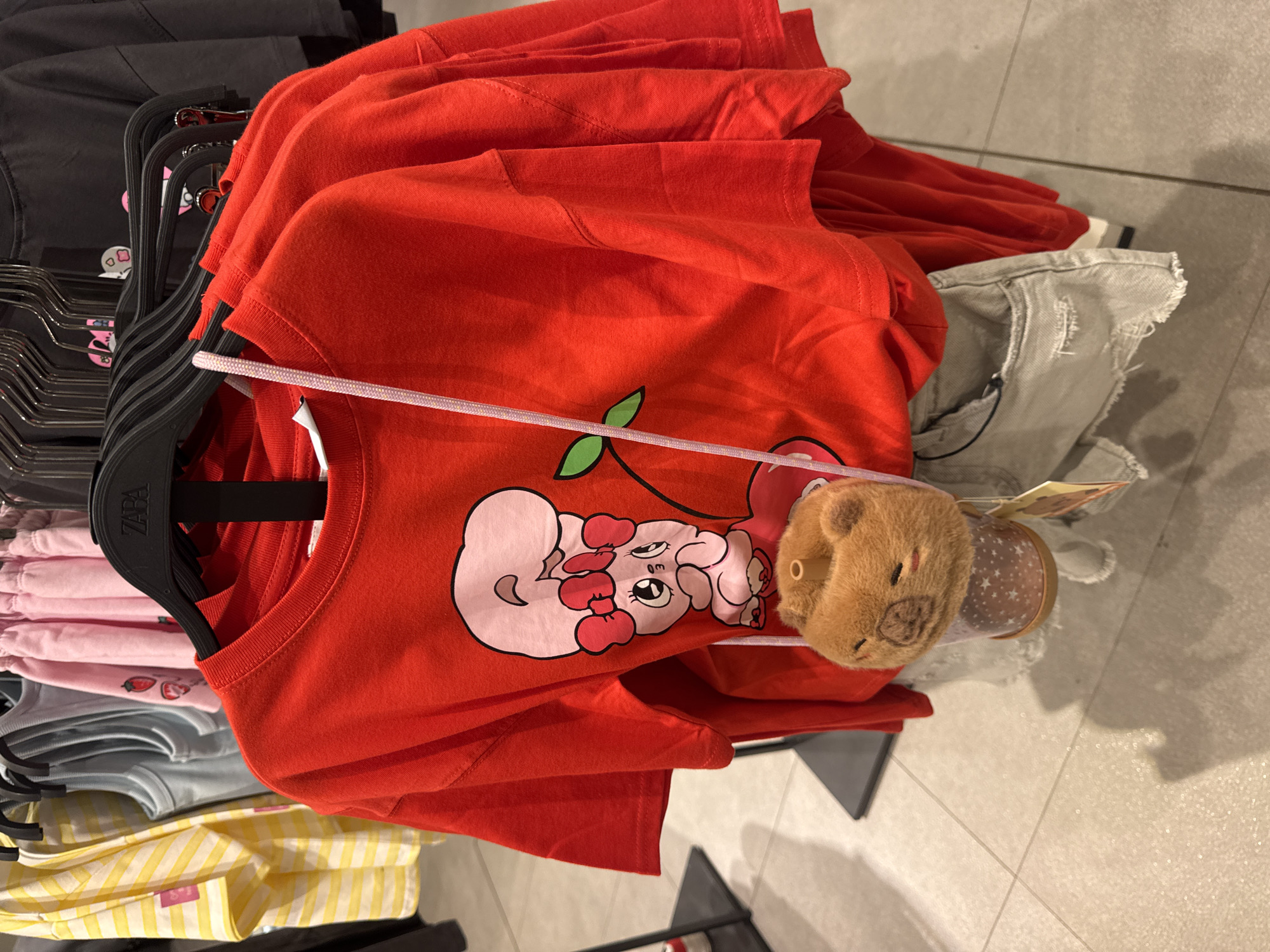 A red t-shirt with a cartoon drawing of a character wearing a large bow and holding a cherry is displayed on a hanger in a clothing store. The t-shirt is paired with light-wash denim shorts. A small, fluffy, bear-shaped purse is attached to the t-shirt by a light pink string. The clothing is hanging on a metal rack in what appears to be a retail environment with tiled flooring. Other t-shirts in various colors and patterns are visible on hangers in the background. The scene is captured in a close-up, frontal view, with the focus on the red t-shirt and its accessories.
