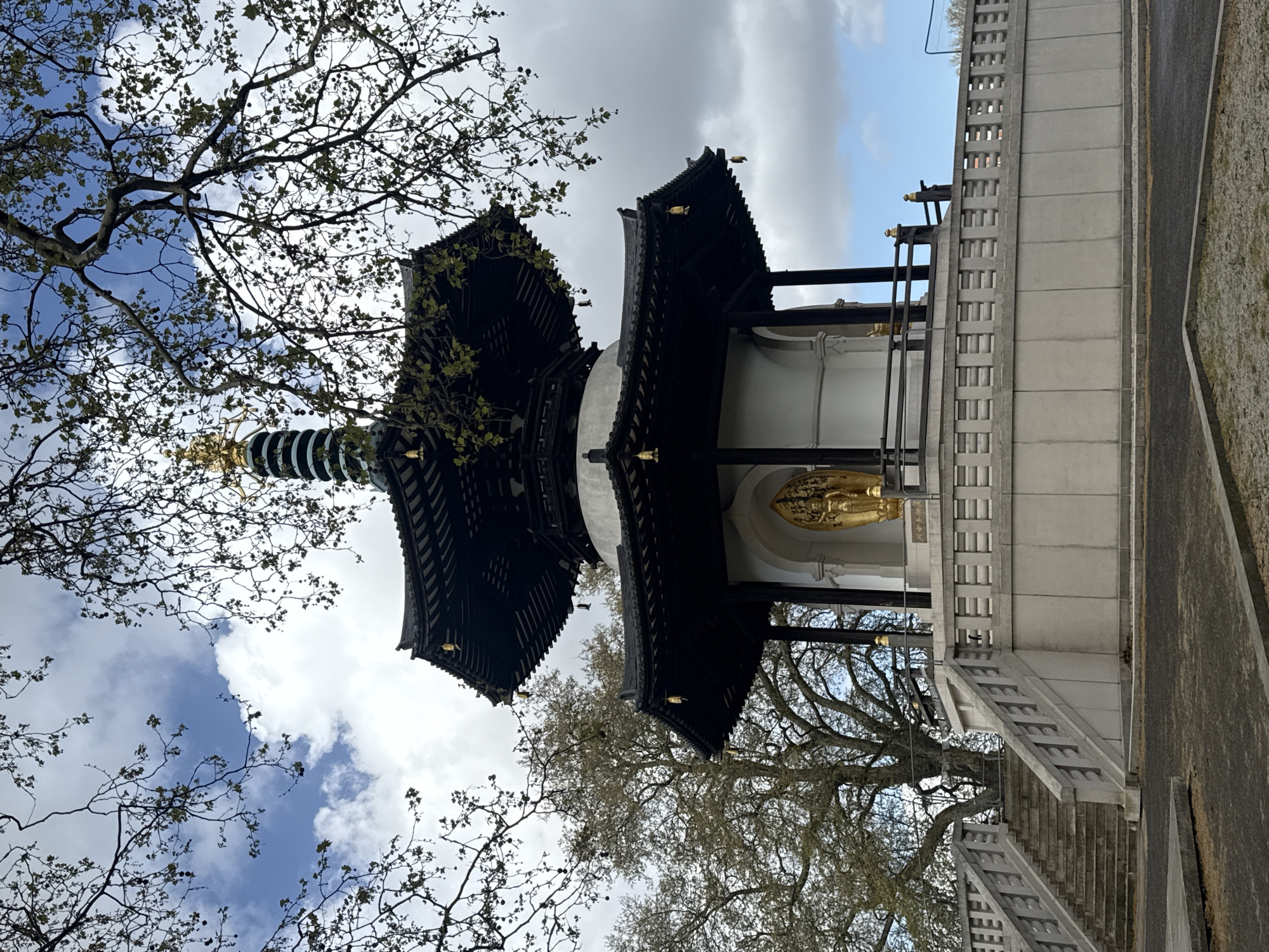 The image shows the Peace Pagoda in Battersea Park, London, United Kingdom. The pagoda is a two-tiered Buddhist structure with a dark-colored roof and a gold statue of Buddha. The pagoda is surrounded by trees and a stone staircase leads up to it. The sky is cloudy with patches of blue.