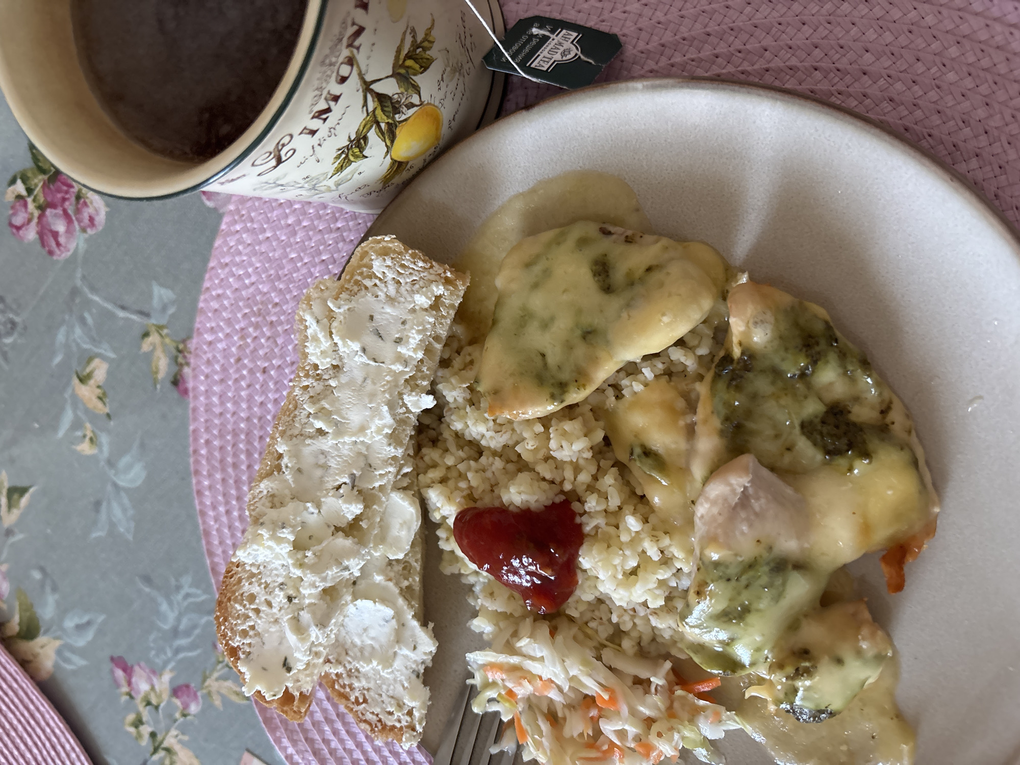 A close-up, overhead shot shows a meal on a plate, set on a pink woven placemat. The meal consists of couscous, coleslaw, and chicken covered in melted cheese and a green sauce, with a dollop of ketchup on the couscous. To the left of the plate, two slices of bread topped with cream cheese are visible. A mug with a floral design, containing a dark liquid and a tea bag, sits to the right of the plate. The background is a tablecloth with a floral pattern. The location is not specified.