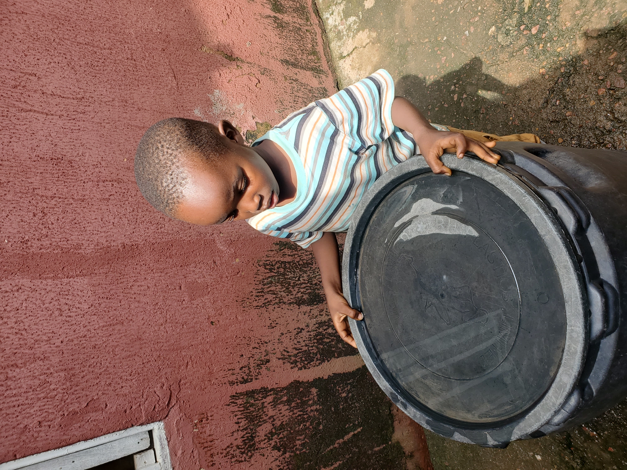 A young boy, appearing to be of African descent, stands outside next to a large black plastic barrel in Jos, Nigeria. The boy is wearing a striped t-shirt and khaki pants. He is looking down at the barrel and has his hands placed on its lid. The barrel is partially full of water, as indicated by the reflection on the lid. The background consists of a textured, reddish-brown wall with some staining and a window frame on the left side. The ground is composed of dirt and small stones, with some damp patches suggesting recent moisture. The lighting indicates it is daytime, casting a shadow of the boy onto the ground.