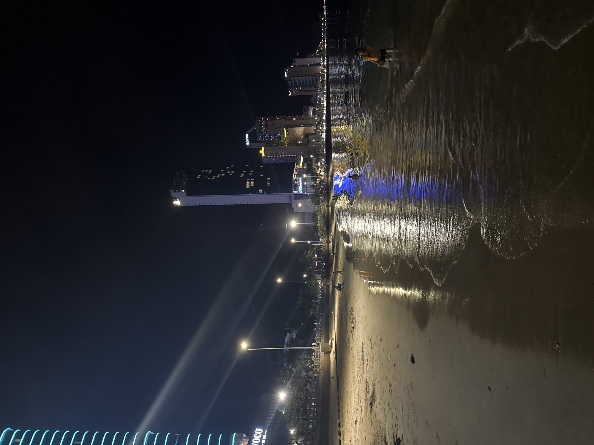 The image captures a nighttime scene on a sandy beach in Bac Nha Trang Ward, Vietnam, illuminated by artificial lights from the city skyline and street lamps. In the foreground, gentle waves lap onto the shore, reflecting the lights from the buildings and creating a shimmering effect on the wet sand. Two children are visible in the shallow water; one in the mid-ground appears to be playing, and another in the right foreground is wading.
Across the beach, a paved walkway is lined with trees and more street lamps. A prominent building on the far left features a vertical blue light display with the text "VOCO" visible. Behind the walkway, a cluster of high-rise buildings forms the city skyline, some with illuminated windows and colorful lighting. The overall atmosphere is one of urban activity extending to the coastline at night.