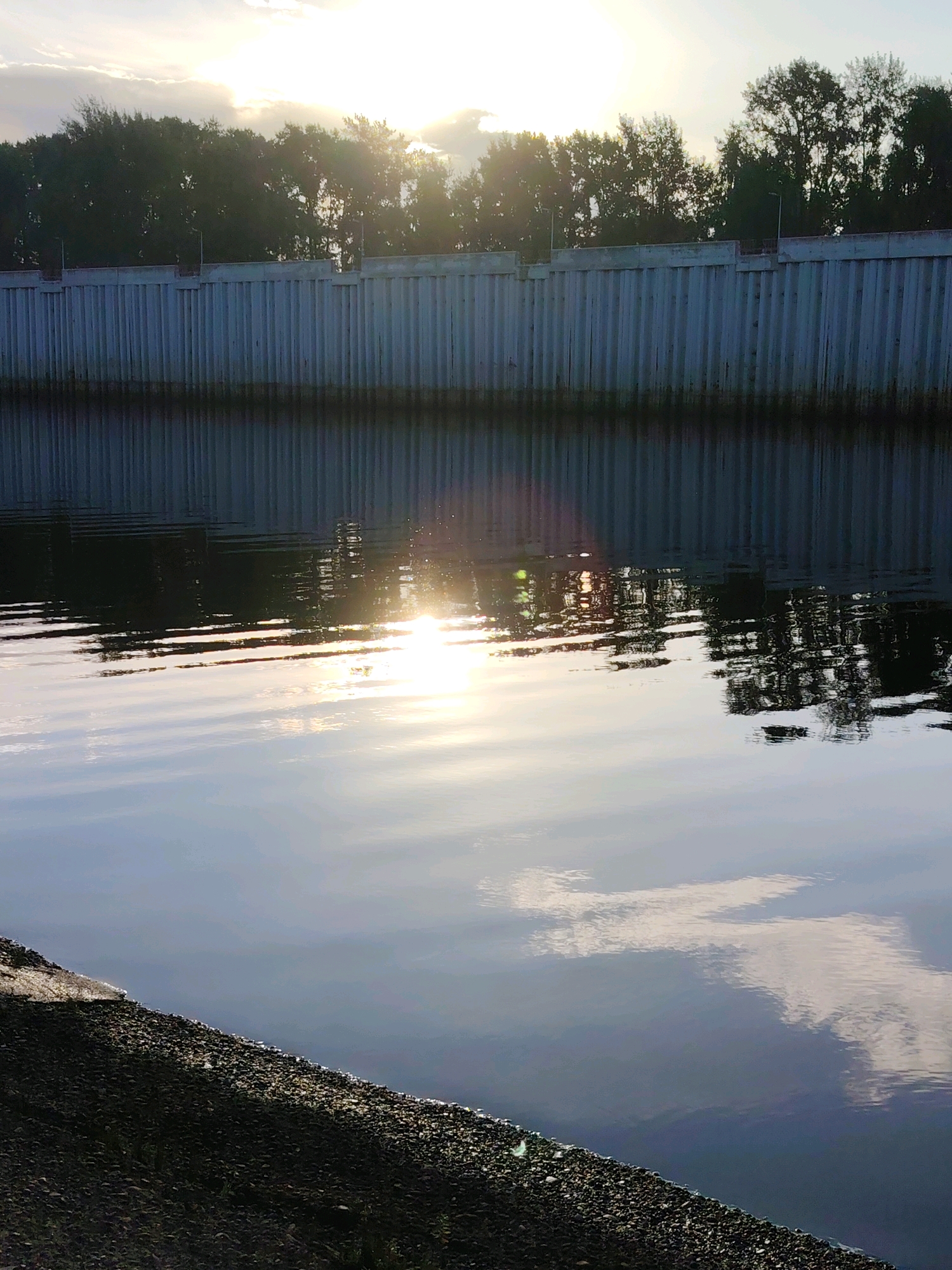 The image depicts a calm body of water, likely a river or canal, reflecting the sky and trees. The sun is low in the sky, creating a bright glare on the water's surface. A long, grey, corrugated metal wall runs along the edge of the water, with trees visible behind it. The foreground shows a stony bank sloping down to the water. The scene is likely located in the city of Chaikovsky, Russia, given the presence of such industrial-style walls often found along waterways in the region, and the general atmosphere of a northern European landscape. There are no people or animals visible in the image, and no actions or interactions are taking place.