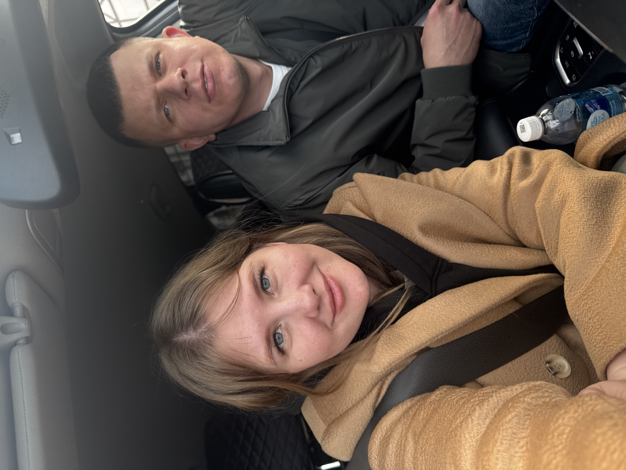 This image captures two individuals inside a car, likely a selfie taken during the daytime. Both subjects have light skin and light blue eyes, and are looking towards the camera with gentle smiles.
In the background, a man with short, dark hair is wearing a dark jacket over a white t-shirt. He appears relaxed, seated in the vehicle.
In the foreground, a woman with long, light brown hair is wearing a warm, light brown or camel-colored coat over a dark top, with a black seatbelt fastened across her.
The setting is clearly the interior of a car, with elements such as an overhead console, seatbelts, and part of a car seat visible. A plastic water bottle with a white cap and a blue label is positioned near the man. The label clearly shows Russian text, "АРИНА РОДНИКОВАЯ," indicating "Arina Spring Water." The overall atmosphere is casual and pleasant. The location context for this image is Ферма, Russia.