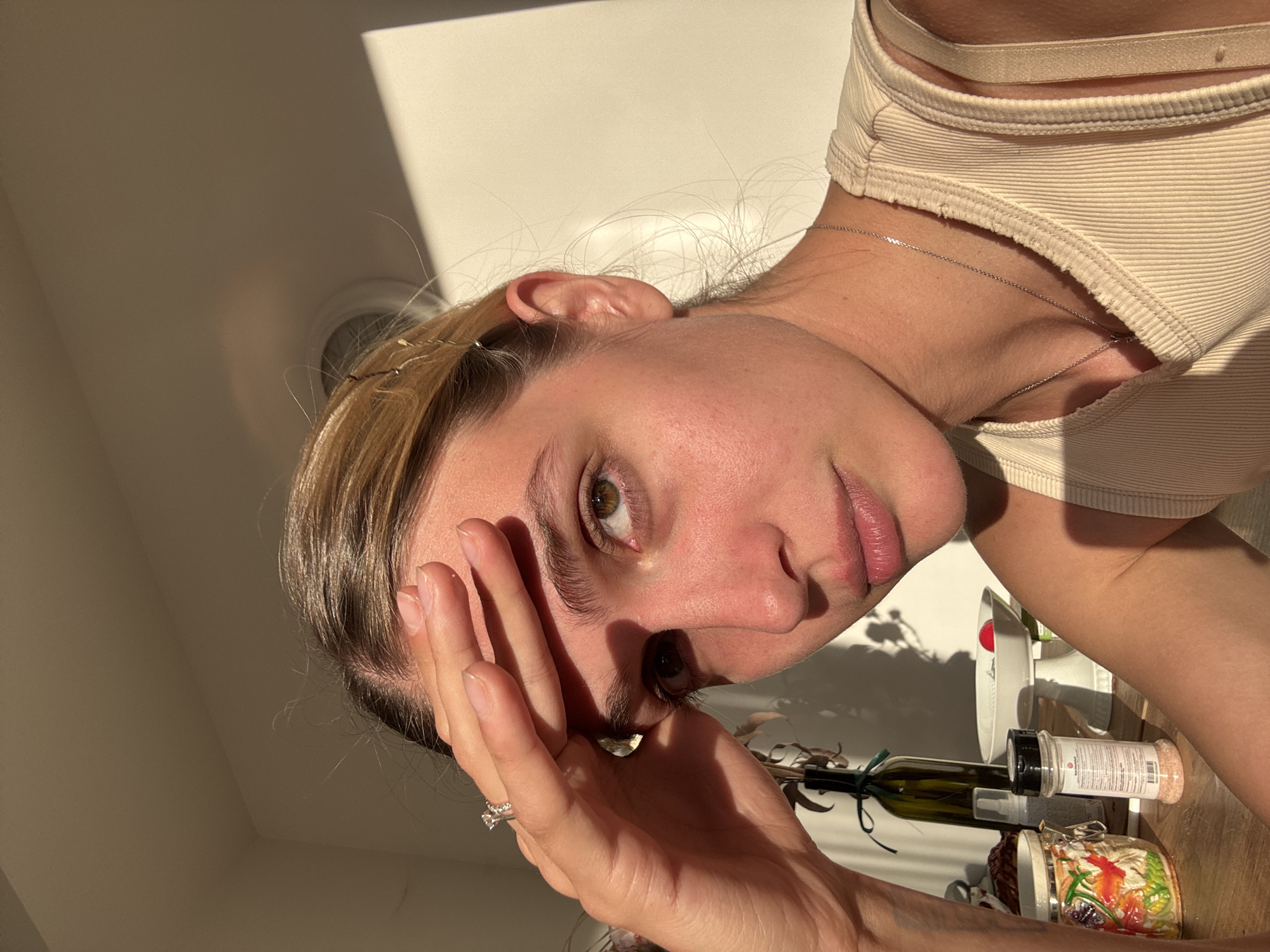 A woman is positioned at an oblique angle, looking directly at the camera with her hand touching her forehead. She has fair skin, light brown hair, and is wearing a light-colored ribbed tank top with thin straps and a silver necklace. Her hair is parted on the side, with some strands pulled back. A ring is visible on her hand.
Behind the woman are various objects on a wooden surface: a small green bottle tied with black twine, a decorative jar, and two spice containers. There is also a white object that appears to be kitchen equipment. Shadows are cast on the background wall and the objects on the surface. The scene is lit with natural sunlight. The location is unspecified, only identified as Unknown, Unknown.