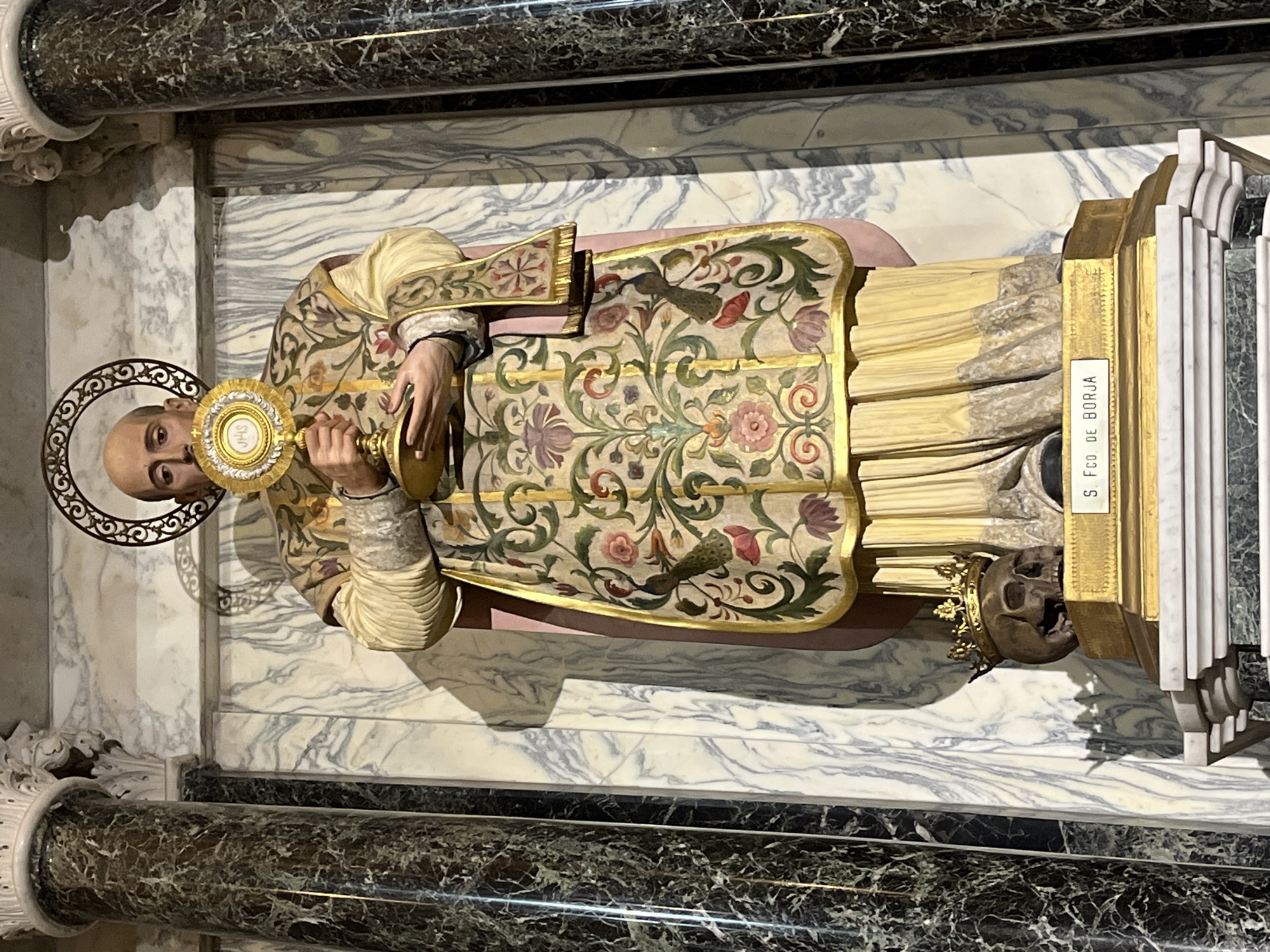 The image shows a statue of Saint Francis Borgia, also known as San Francisco de Borja, located in the Iglesia del Santísimo Sacramento in Buenos Aires, Argentina. The statue depicts the saint recumbent, dressed in ornate vestments. He is holding a monstrance. A skull topped with a golden crown lies at his feet. The statue rests on a marble shelf. Above the statue is marble paneling. Below the statue is a column with decorative molding.