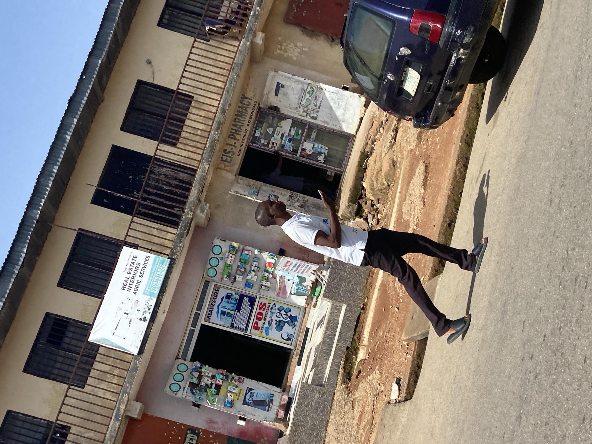The image captures a street scene in Jos, Nigeria, under a clear blue sky. A man wearing a white polo shirt and dark trousers is walking on the paved road, holding a mobile phone in his right hand and looking at it. He is wearing black sandals. Behind him, a dark blue station wagon is parked, showing some wear and tear on its rear bumper. The road is flanked by a dirt and rock embankment to the left and a weathered building with multiple shops and balconies to the right.
The building's facade features shops with open entrances and signage. One sign indicates "REAL ESTATE" with a depiction of houses and services like "RENTING & REMODELLING" and "BUYING & SELLING". Another shop is labeled "POS & PHONE ACCESSORIES", displaying various small items on its shelves. Above these shops, a balcony with a railing is visible. A banner is stretched across the balcony railing, advertising "REAL ESTATE INTERIORS AGRIC. SERVICES" with contact phone numbers. Further to the right, a shop with the name "REIS-J. PHARMACY" is identified, also displaying phone numbers and a P.O. Box address. Items are visible on shelves inside the pharmacy. The building's exterior shows signs of age and wear, with some peeling paint and exposed concrete. The overall environment suggests a commercial area within a city.