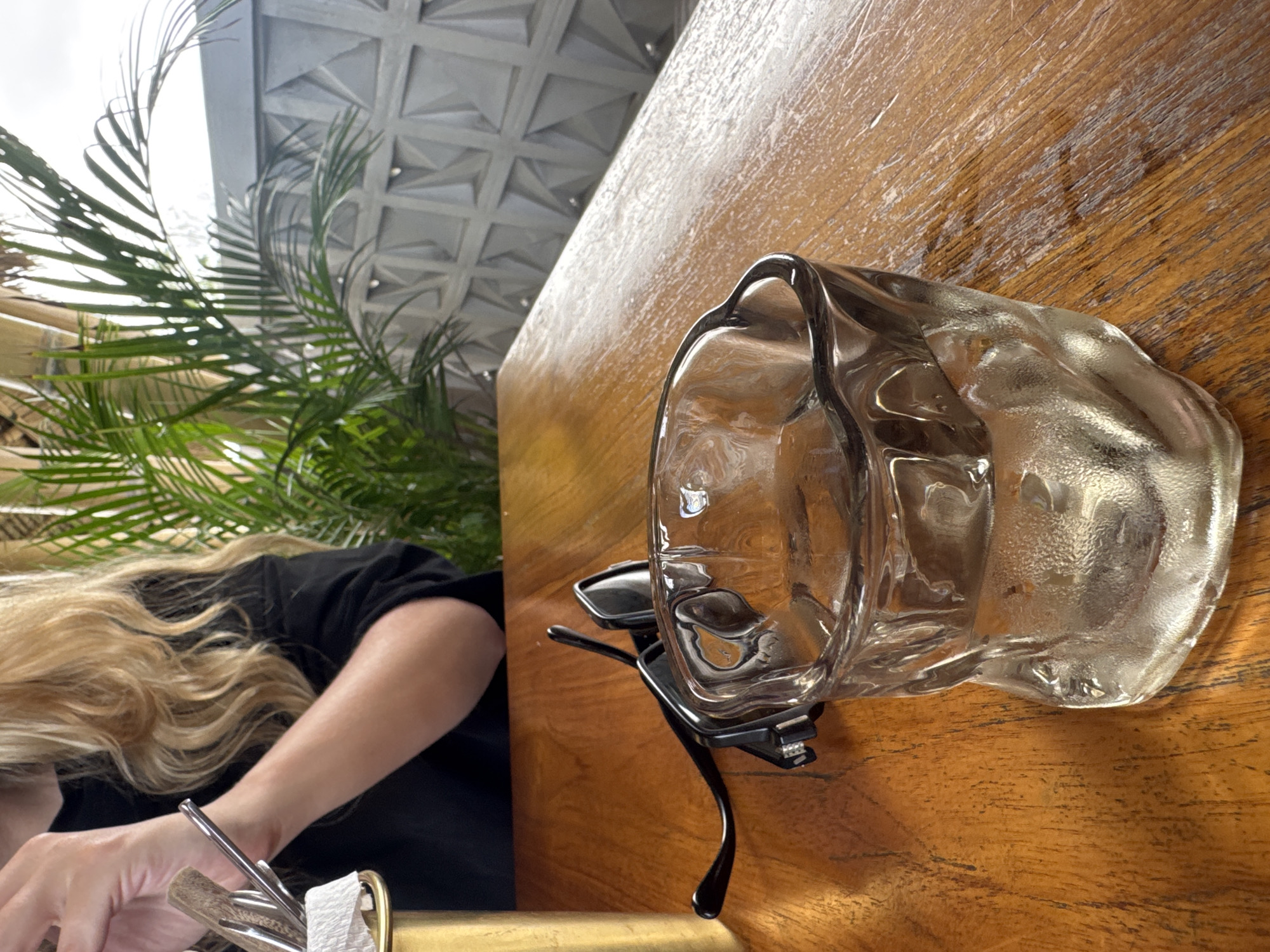 The photograph captures a scene at a dining table, likely indoors, with a woman seated on the left. She has long blonde hair and is wearing a black top. Her hands are visible as she interacts with silverware near a partially visible white napkin.
On the right side of the table, which has a light brown wood grain finish, sits a clear glass. A pair of black sunglasses is positioned beneath the glass. A potted green plant is visible behind the woman. The background shows a ceiling with a patterned, possibly geometric, design. The setting is not explicitly stated to be in Kuta Selatan, Indonesia, but it might be a location within the area.