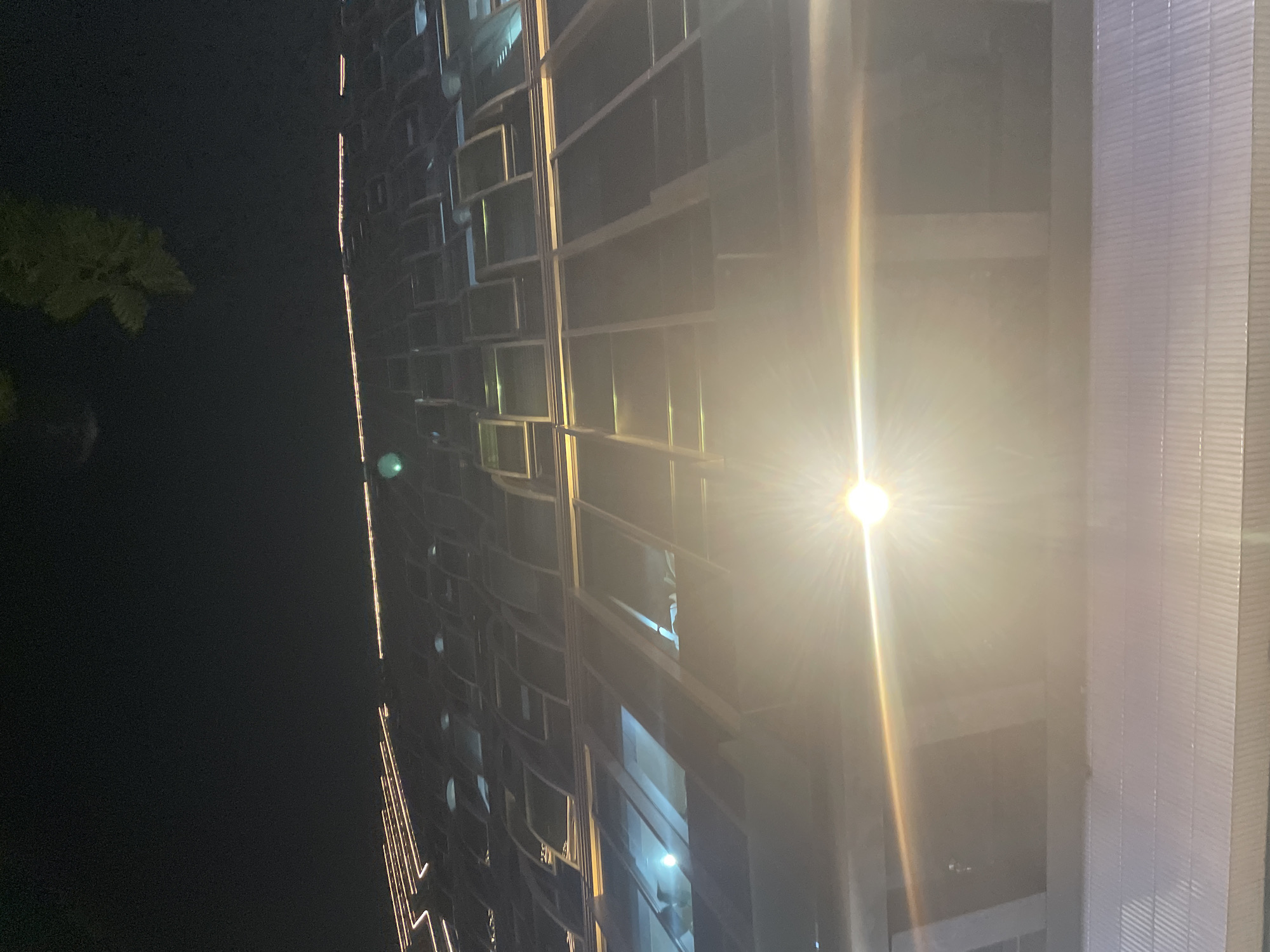 This is a nighttime photograph of a modern building in Thủ Đức, Vietnam. The building features a facade of repeating rectangular windows with thin, light-colored frames. A bright, starburst-shaped light source, possibly a streetlamp or a strong reflection, dominates the right side of the image, casting a warm glow across the building's surface and creating lens flare.
The building's design suggests a residential or office complex, with numerous illuminated windows visible, hinting at activity within. Some windows appear dark, while others show faint interior lights. A vertical line of smaller lights runs along the left edge of the building, adding to its illuminated appearance.
To the left of the building, a dark, silhouetted tree with sparse foliage is visible against the night sky. The overall scene is one of urban nighttime, characterized by artificial lighting and the stark contrast between the dark sky and the illuminated structure. The weather appears clear, as there are no visible signs of rain or fog. No people are clearly discernible, though indistinct shapes might be present within the lit windows. There is no visible text in the image.