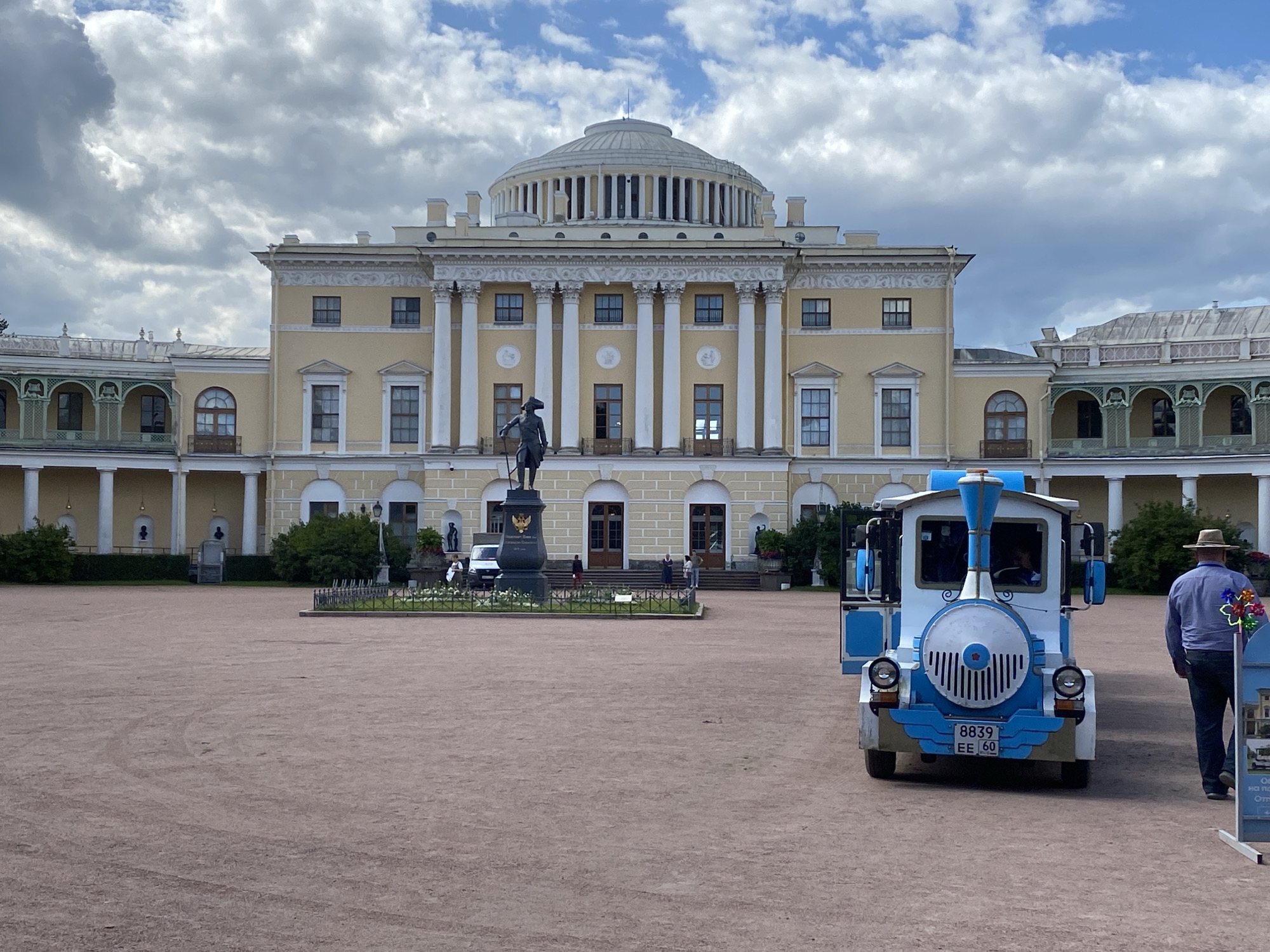 The image shows the Pavlovsk Palace in Pavlovsk, Russia, a neoclassical palace located south of Saint Petersburg. The palace is a large, yellow building with a prominent dome and white columns. In the foreground, there is a small, blue and white tourist train, resembling a cartoonish locomotive. A man wearing a hat and a blue shirt is walking away from the train towards the right side of the frame. The ground in front of the palace is a reddish-brown gravel, and there is a statue of a man on horseback in the middle ground. The sky is cloudy with patches of blue.