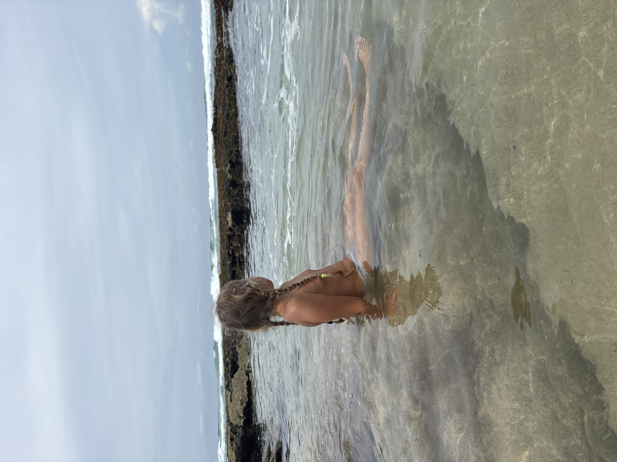 A young person wades in the shallows of a body of water near the shore at Thalaramba, Sri Lanka. The individual is facing away from the camera, and only their back, shoulders, and arms are visible above the waterline. The person has light-colored hair in braids. The water is clear and reflects sunlight. Rocks and waves appear on the left side of the frame. The sky is overcast.