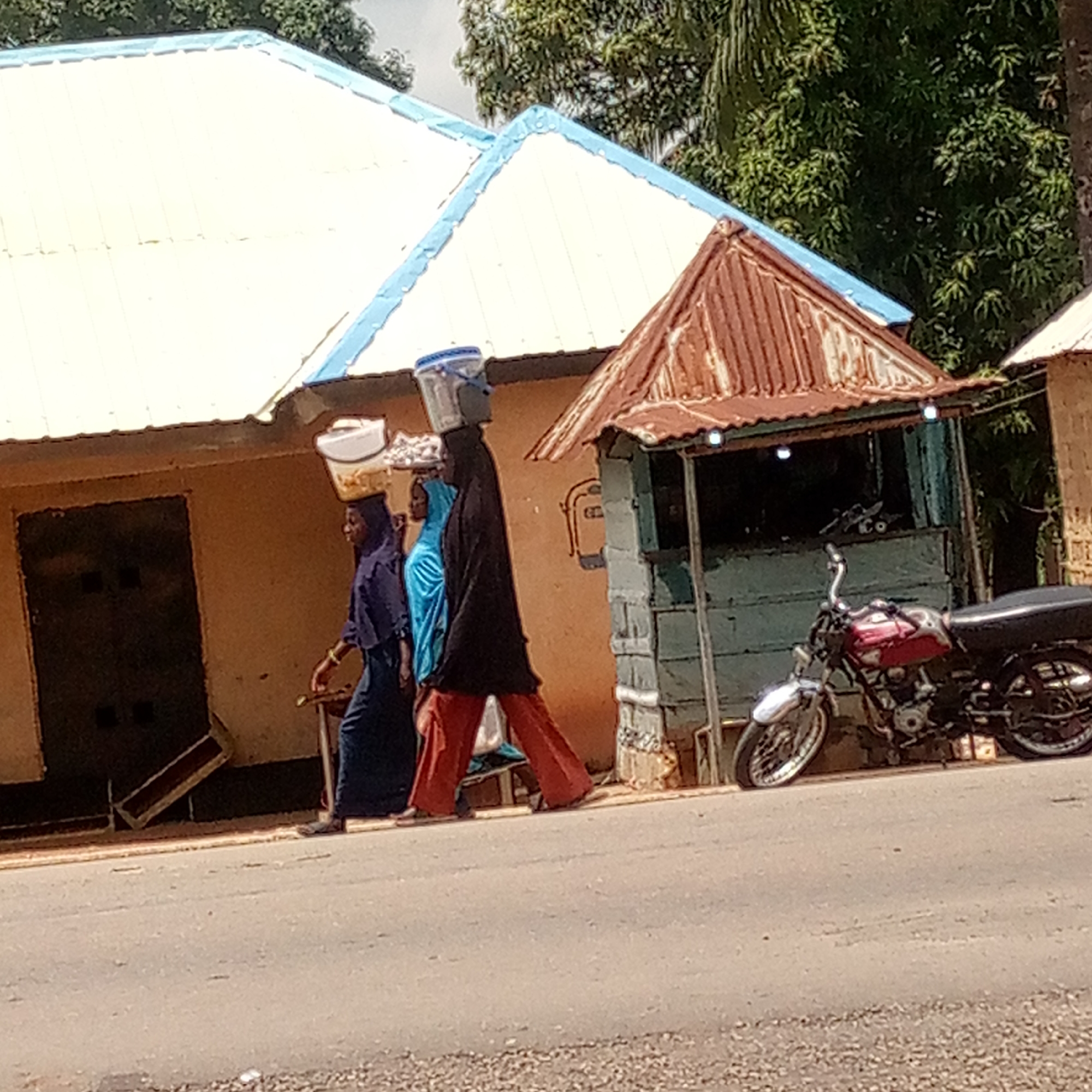 The image captures a vibrant street scene, likely in Mararaba, Nigeria, under bright daylight. Three women are walking from left to right along the side of a paved road. Two of them are skillfully balancing items on their heads, a common method of transport in the region.
The lead woman, dressed in a black abaya and reddish-brown trousers, carries a white and blue bucket on her head. Behind her, another woman, wearing a light blue dress or hijab, balances a white bowl or container. The third woman, further back and partially obscured, is also carrying a white container on her head while dressed in a dark blue outfit.
To the left, a building with light brown walls and a distinctive white corrugated iron roof with blue trim provides the backdrop. It features a dark, recessed doorway. Adjacent to it, on the right side of the frame, stands a smaller, rustic structure, possibly a kiosk or small shop, with a rusty brown corrugated iron roof and faded blue-grey walls. Small lights are strung across its front. A red and black motorcycle is parked in front of this smaller building.
The road is asphalt, and the shoulder where the women are walking appears to be a mix of dirt and gravel. Lush green trees are visible in the background under a bright, clear sky, indicating sunny weather. No discernible text is visible on any of the objects or buildings. The scene depicts daily life and common modes of transport in a local community.