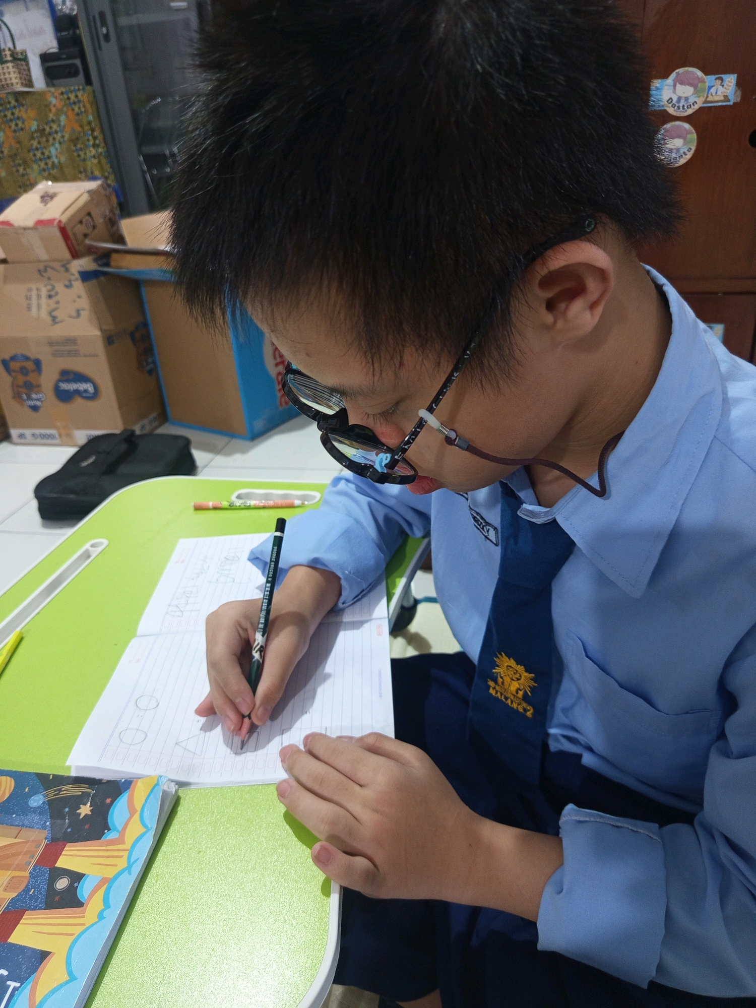 The image depicts a young person, likely a student, wearing a light blue collared shirt and a dark blue tie with an emblem that appears to say "MALANG 2". They are seated at a bright green table, intently writing in a notebook with a pencil. The notebook is open to a page with hand-drawn shapes, including circles and a triangle. The person is wearing glasses with a black frame and a thin brown cord. In the background, there are several cardboard boxes, one of which has writing on it, and a black bag is visible. There are also some decorative stickers on a wooden surface behind the student. The setting appears to be indoors, possibly a home or a school environment, with the presence of school-related materials. The style of the uniform and the inscription on the tie suggest a connection to Malang, Indonesia.